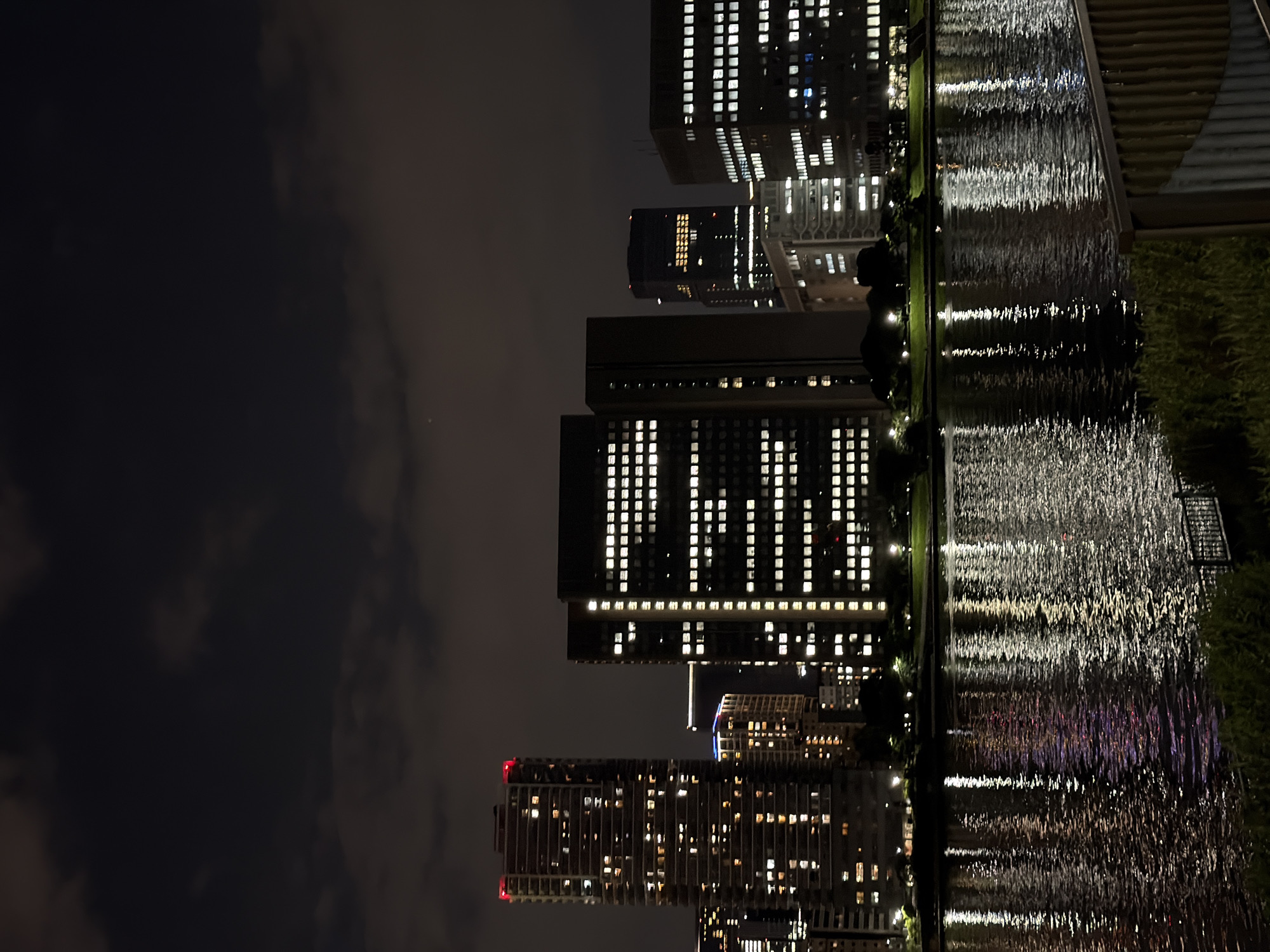 A nighttime cityscape in Koto, Japan, is captured from across a body of water. Tall, illuminated buildings dominate the background, their windows glowing with a warm, white light. A prominent skyscraper on the left features a red illuminated top. A building in the center has a pattern of illuminated squares forming a grid-like design. The water in the foreground reflects the lights of the city, creating shimmering streaks of color. The sky above is dark with scattered clouds. The foreground includes a dark, grassy embankment and a portion of a wooden railing. There are no visible people or animals in the scene.