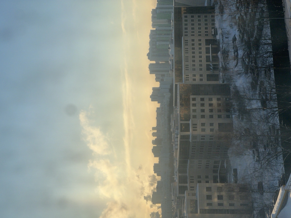 A cityscape under a sky filled with soft, pastel-colored clouds. Tall, multi-story buildings with repetitive window patterns dominate the scene. Bare trees stand alongside the buildings, with a dusting of snow visible. The composition suggests a winter setting with an atmosphere of calm and muted tones.