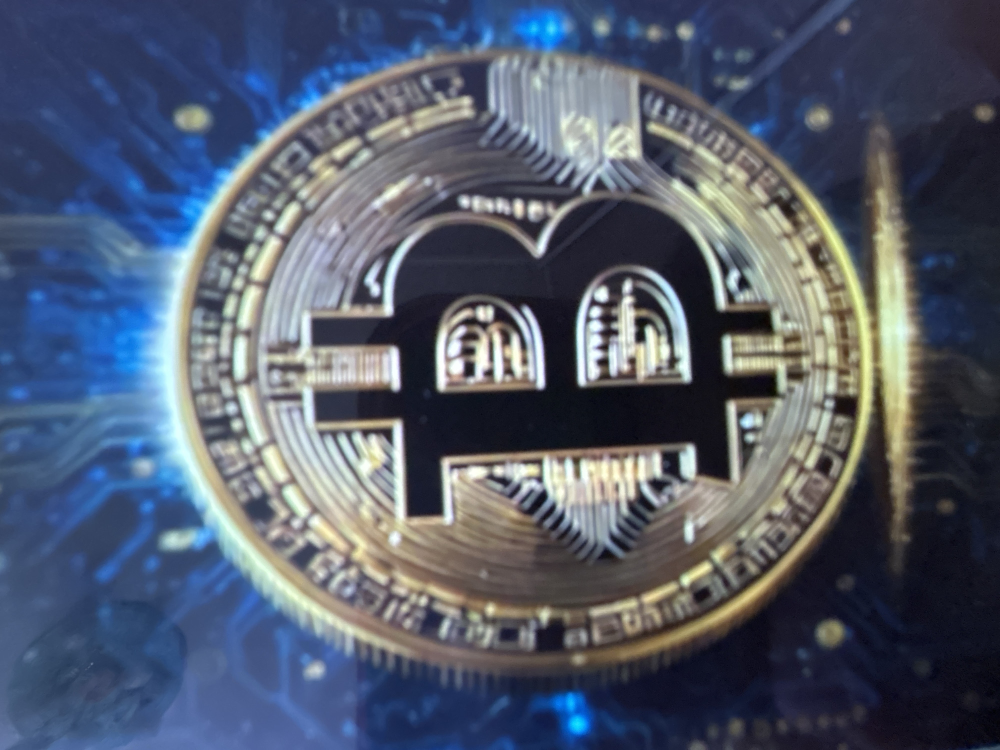 The image displays a close-up, slightly blurred view of a golden Bitcoin coin against a dark, abstract background with blue circuit-like patterns and glowing lights. The coin is centrally positioned and illuminated, showcasing its metallic sheen and intricate design, including the iconic "B" symbol. The background suggests a digital or technological theme. There are no discernible elements in the image that could be used to identify a specific location in Gelendzhik, Russia, nor are there any people, actions, or interactions depicted.