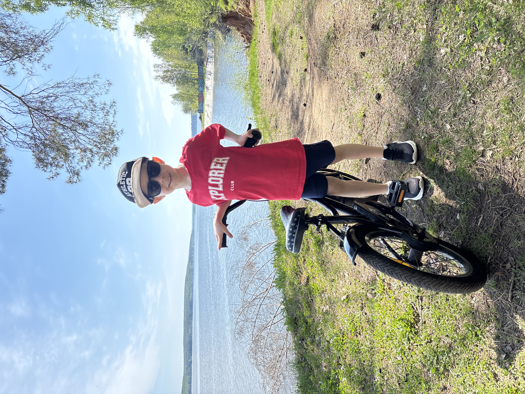 A young boy wearing a red t-shirt with the word "EXPLORER" and the word "CLUB" printed on it, dark shorts, a baseball cap, and sunglasses is standing on a black bicycle on a grassy path near a body of water. The path is edged with trees. A concrete structure is visible across the water. The location is likely near Chaikovsky, Russia.