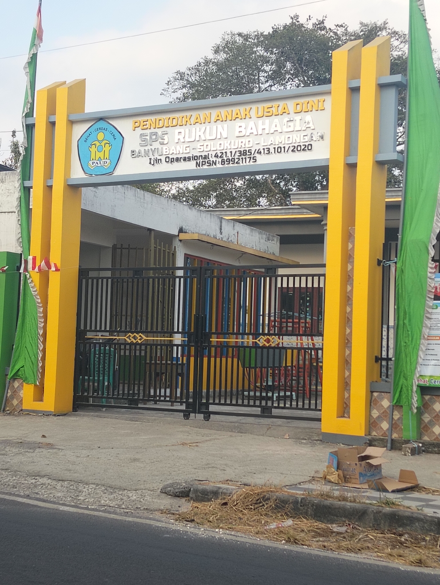 This is a full shot, eye-level outdoor photograph of the entrance to a school building. The entrance is flanked by two tall, bright yellow pillars. A black metal gate with decorative elements stands between the pillars. The gate is partially open, revealing a building with a yellow roofline. Colorful vertical stripes of red, blue, and yellow adorn the front of the building behind the gate. A sign with white text on a grey background is mounted on the structure between the pillars. The sign reads "PENDIDIKAN ANAK USIA DINI SPS RUKUN BAHAGIA" and includes address information and an operational permit number. A green flag hangs to the right of the entrance, and a smaller flag with red and white stripes is attached to the left pillar. The ground in front of the school is paved, with a curb and a grassy area along the roadside. Discarded cardboard boxes and dry grass are visible on the sidewalk. The sky is overcast. The overall impression is of a public building in an Indonesian town.
