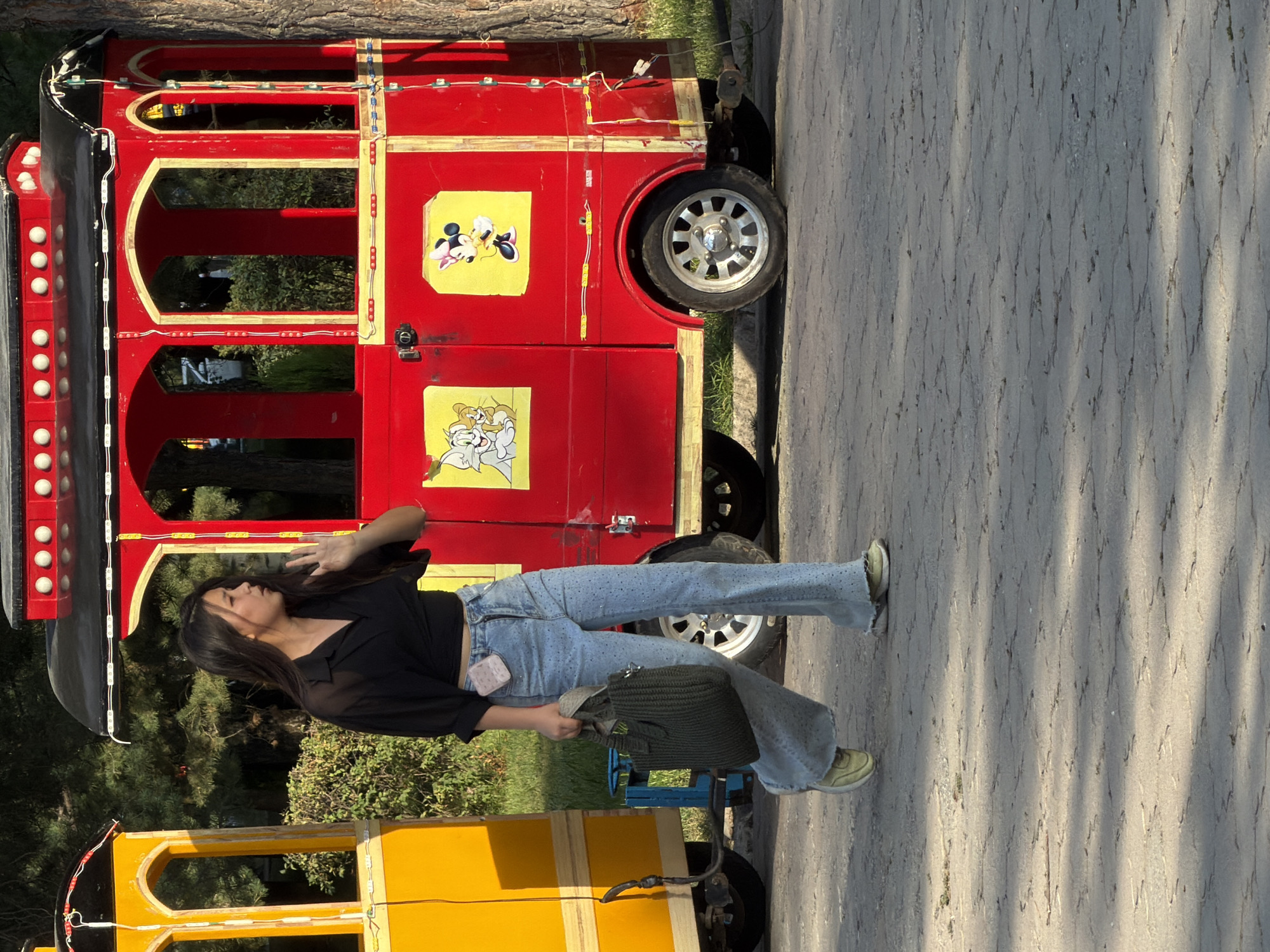 A young woman with long dark hair is walking in front of a red train car attraction. She is wearing a black off-the-shoulder top, light blue jeans embellished with rhinestones, and yellow sneakers. She is carrying a dark green woven tote bag in her left hand. Her right hand is raised to her face, and she appears to be speaking. The train car has windows and is decorated with images of cartoon characters, including Tom and Jerry, and Mickey Mouse. A yellow train car is partially visible to the left of the red one. The ground is paved with interlocking grey stones, and trees and bushes are visible in the background. The lighting suggests it is daytime.