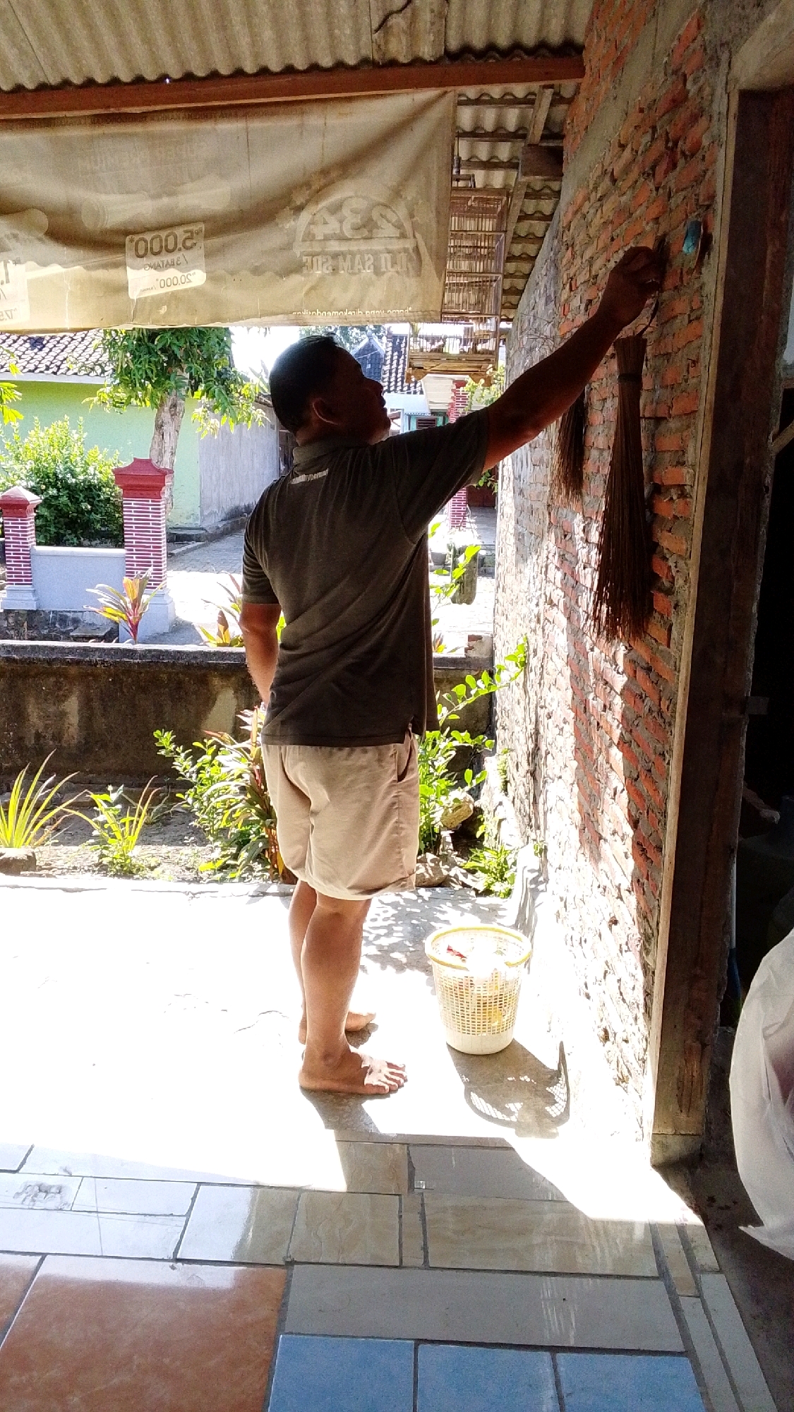 An adult male, wearing a dark grey polo shirt and light tan shorts, stands barefoot outdoors, facing a brick wall. He is holding a broom in his right hand and appears to be cleaning or painting the wall. A white plastic basket filled with items sits on the tiled ground to his left. The scene is illuminated by bright sunlight, casting shadows. In the background, a tiled patio area is visible, along with greenery, a low concrete wall, and ornamental brick pillars. The location appears to be an outdoor area, possibly a patio or entryway, in Lawu, Indonesia, characterized by the architectural style and vegetation.