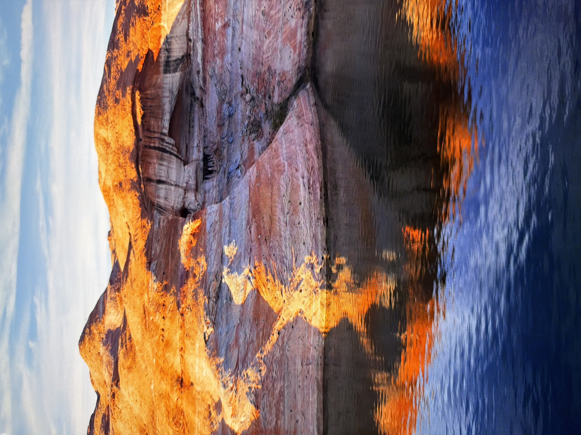 The image depicts a scenic view of a body of water reflecting vibrant orange and purple rock formations under a clear blue sky. The rock formations appear to be layered and eroded, with shadows highlighting their texture and curves. The water is calm, with ripples distorting the reflection of the sky and rocks. There are no people, objects, or actions visible in the scene. The location is not specified as Kyiv, Ukraine, and the landscape suggests a natural desert or canyon environment.