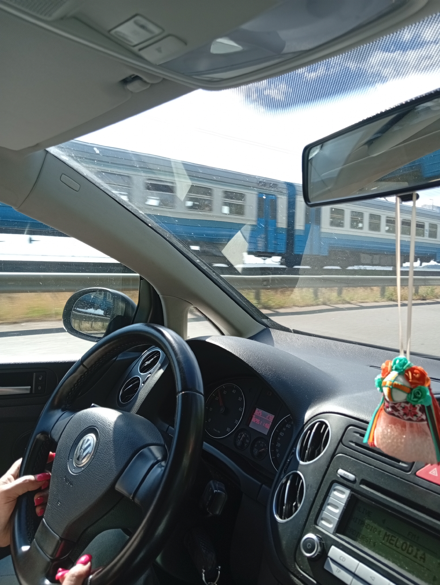 The image shows the interior of a car with a person's hands on the steering wheel, as seen from the driver's perspective. Outside the car, a blue and white train is passing by, blurred due to motion. The car's dashboard is visible, including the steering wheel with a Volkswagen logo, air vents, and a car stereo. A decorative air freshener hangs from the rearview mirror. The scene appears to be a daytime shot, with sunlight illuminating the interior and the exterior. The background suggests an outdoor environment, possibly near a train track within a city.