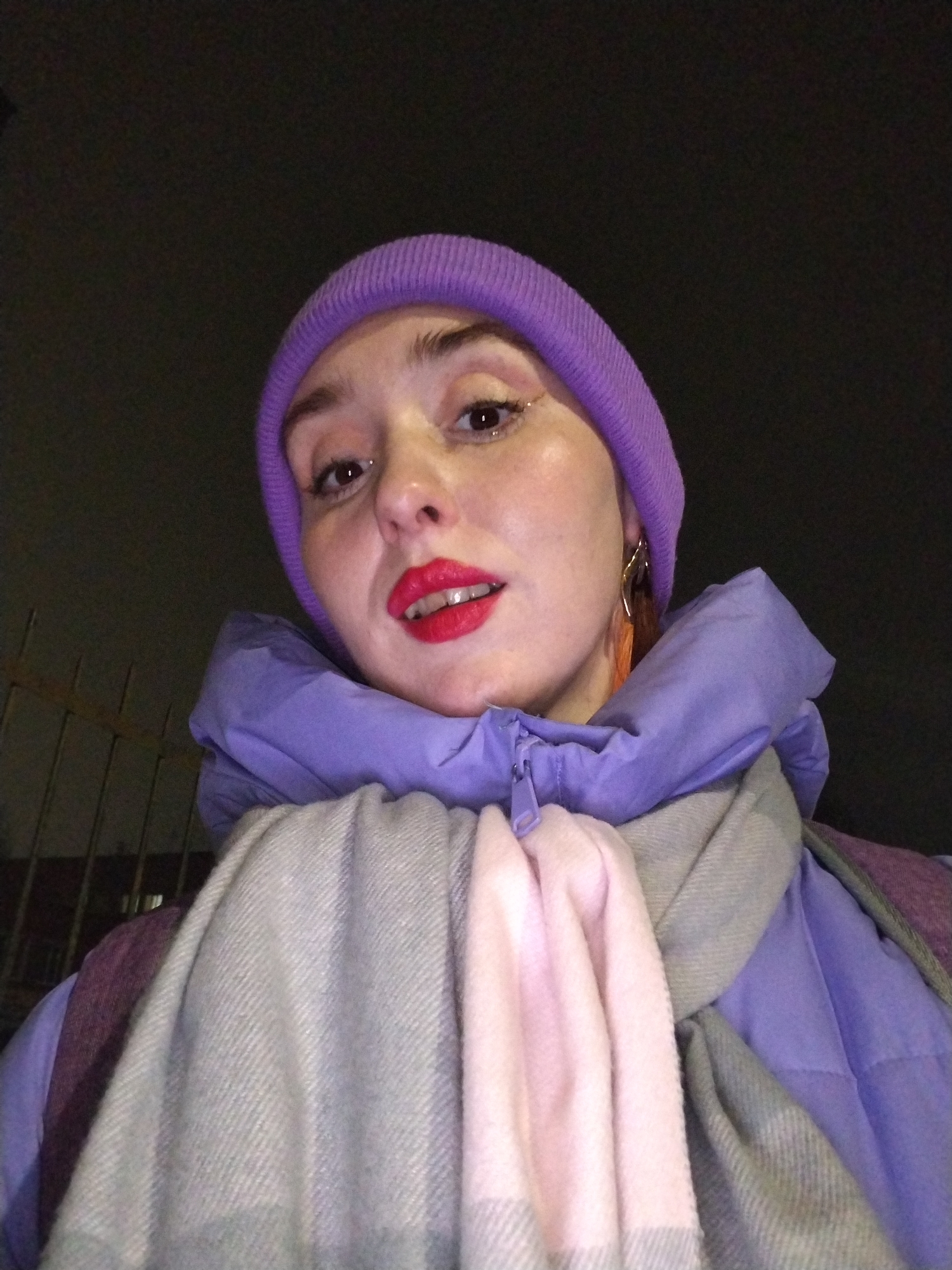 A woman is taking a selfie. She is wearing a purple beanie, a purple puffer jacket with the hood up, and a gray and pink scarf. She has red lipstick and glitter eye makeup. The background is dark. It is difficult to verify the location as Izhevsk, Russia based solely on the image provided. The image does not contain any information that can be used to determine the location.