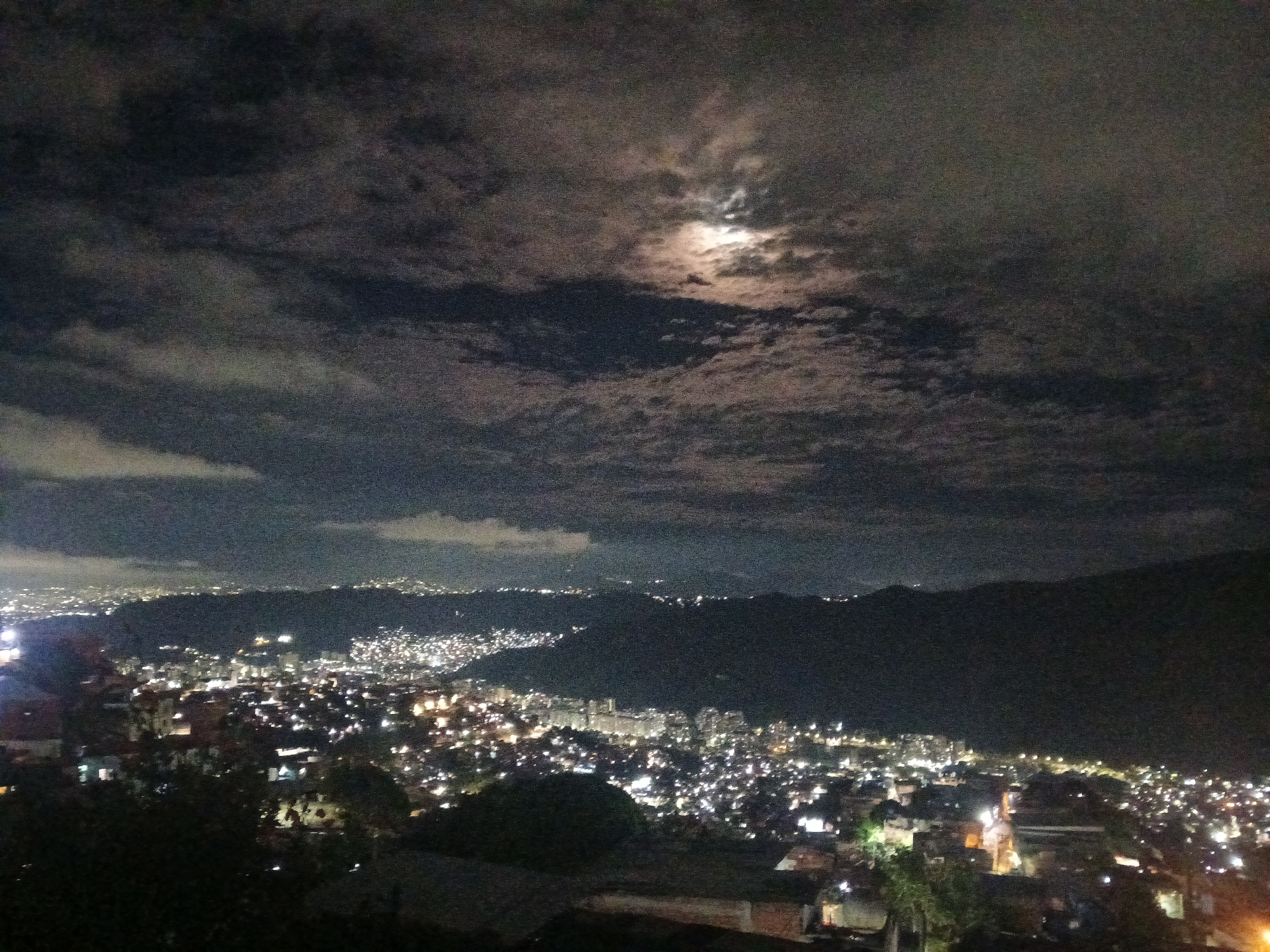 The image shows a night view of Caracas, Venezuela. The city is illuminated with numerous lights, creating a dense urban landscape. A mountain range forms a dark backdrop. The sky is cloudy, and the moon is partially visible through the clouds, casting a faint light. The foreground consists of dark, undefined shapes, possibly trees or buildings.