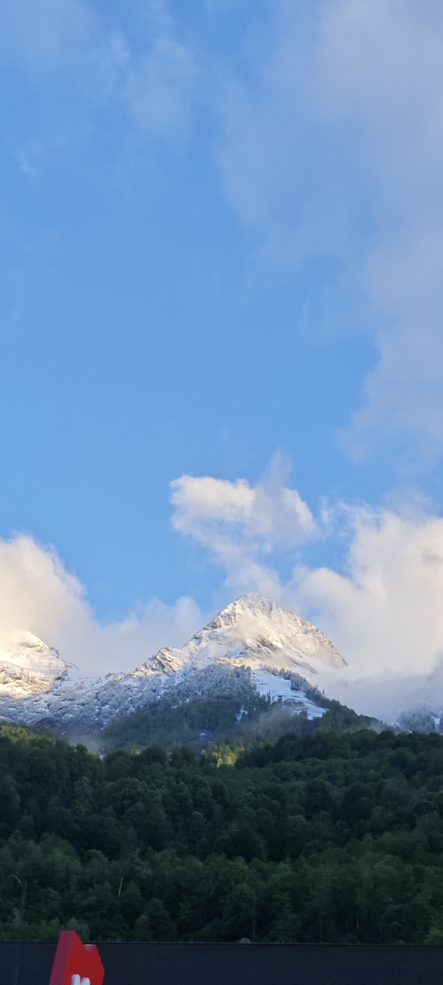 The image presents a mountain range partially covered in snow, with green vegetation at the base. The sky is blue with scattered white clouds. Located in Estosadok, Russia, the foreground contains the tops of trees with a building in the lower foreground with a red and white logo.