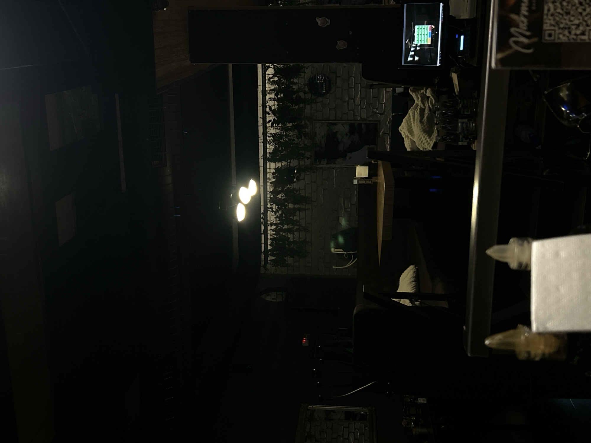 The image shows an interior scene, likely a bar or restaurant, possibly in Plovdiv, Bulgaria. The environment is dark with low lighting, featuring brick walls with greenery and framed art. A computer monitor is visible on a counter, suggesting a point-of-sale system. A metal frame or stand is prominent in the foreground, partially obscuring the view, and it holds a white card. There is no clear indication of people or specific interactions.