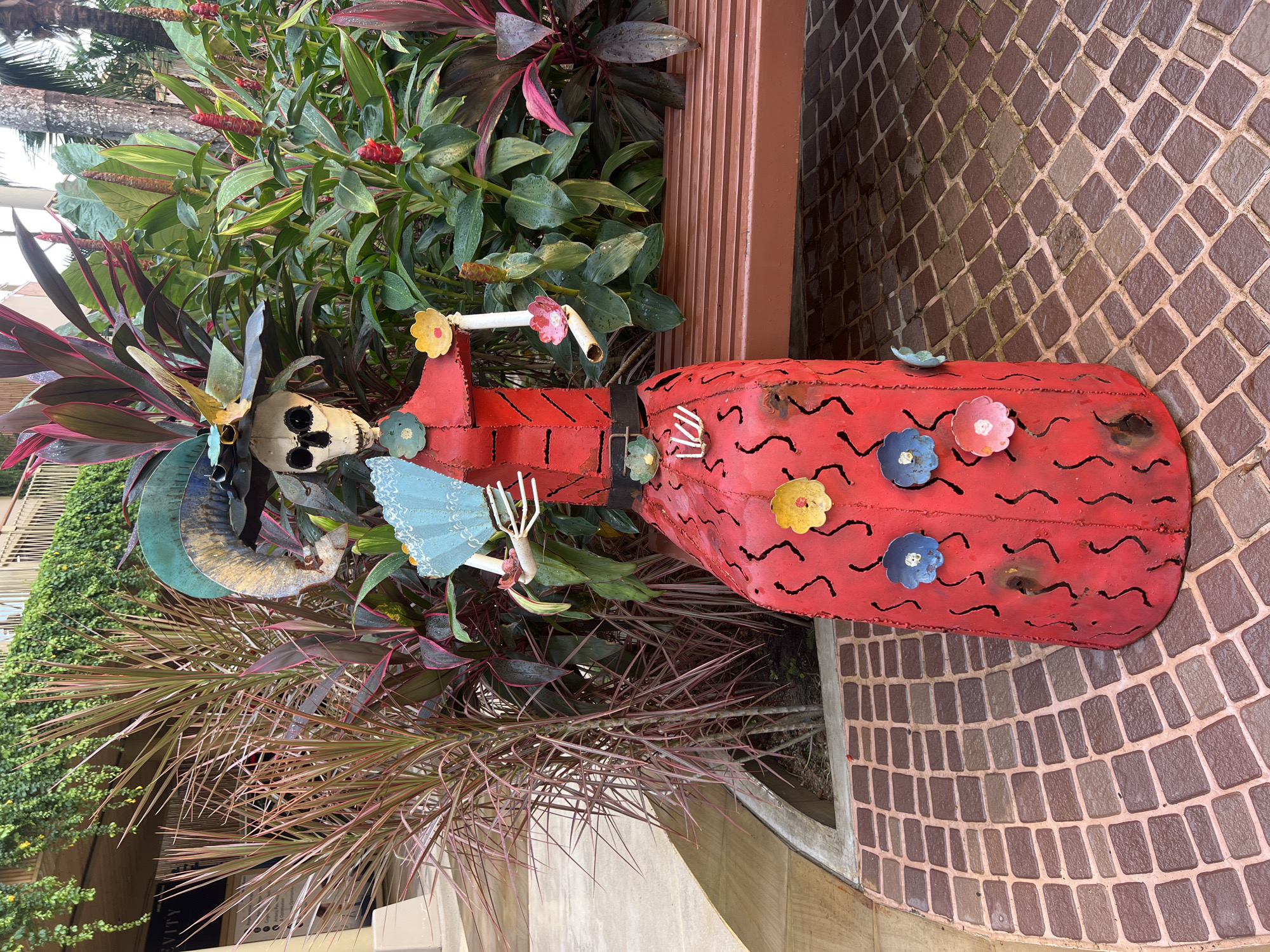 In the city of Choeng Thale, Thailand, a brightly colored, metal sculpture of a skeletal figure in a red dress stands on a brick-patterned walkway. The figure, resembling a traditional "Día de los Muertos" character, wears an ornate hat adorned with a teal feather and yellow floral accents. Its arms are extended, one holding a blue fan, and the other grasping a white flower. The dress is vibrant red, decorated with black wavy lines and various small, colorful flowers—blue, yellow, pink, and green. Behind the sculpture, lush green foliage with red and pink accents thrives, contrasting with a brown wooden bench. The background reveals a building with a balcony draped in greenery. The scene appears to be outdoors, possibly in a garden or courtyard area, with a slightly damp walkway suggesting recent rain.