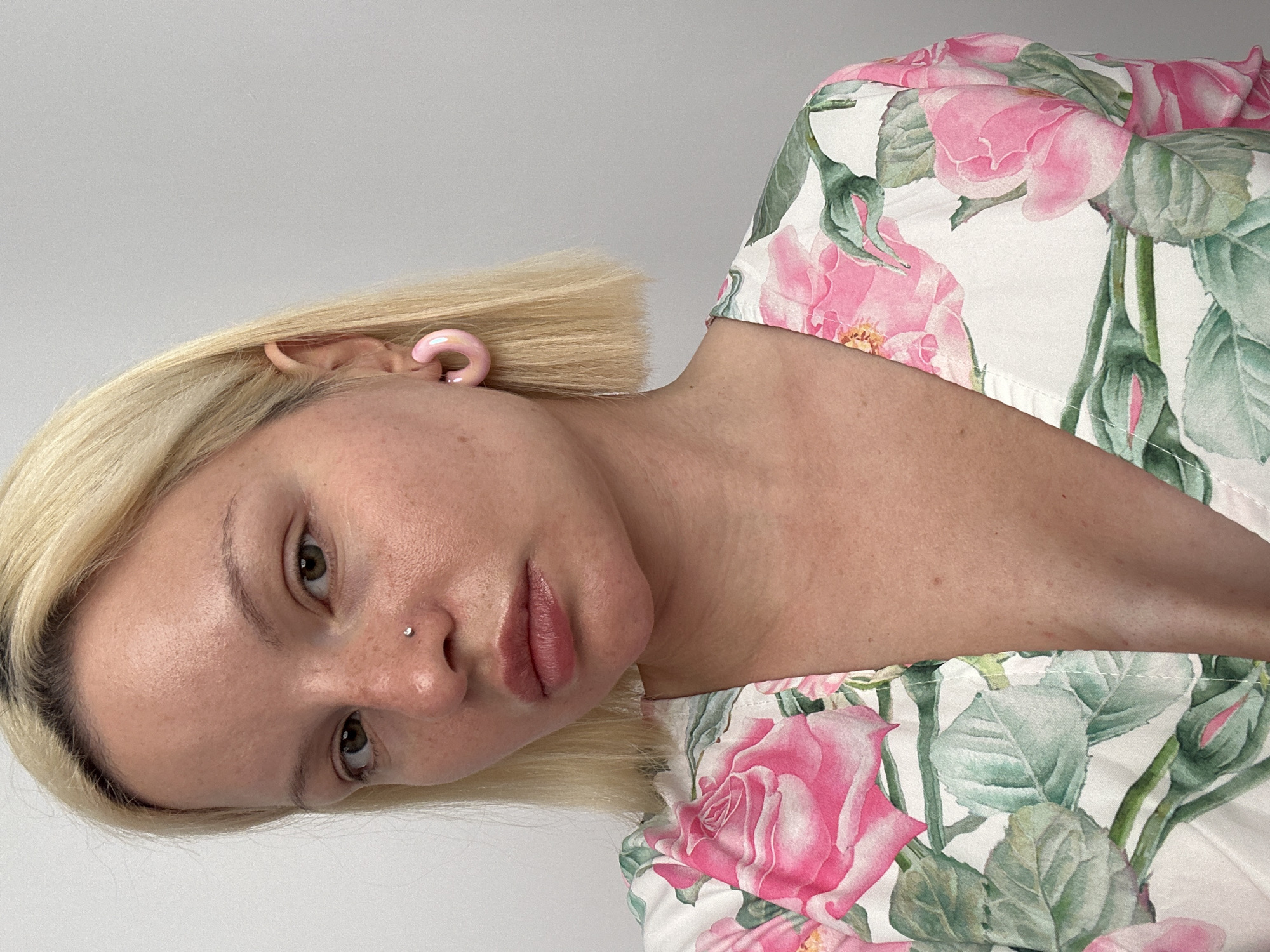 This is a close-up, eye-level portrait of a person, likely a woman, with short blonde hair and fair skin, depicted from the chest up. Her head is tilted slightly to her left, and she looks directly towards the viewer with a neutral to slightly pensive expression.
She is wearing a white garment patterned with large, detailed pink roses and green leaves, suggestive of a floral print top or dress. Her features include light-colored eyes (appearing greenish-hazel), a small, sparkling stud piercing on her right nostril, and a glossy, light pink ear gauge or tunnel visible in her right earlobe, which is partially covered by her hair. Her lips are full with a natural, muted pink tone. Her skin has a natural appearance, showing some subtle freckles.
The background is a plain, light grey or white wall, indicating an indoor setting with clean, soft lighting. The focus is entirely on the subject, with no other objects or individuals present in the frame. The provided context states the location is Budva, Montenegro, though this is not visually evident in the image. No text is visible.
