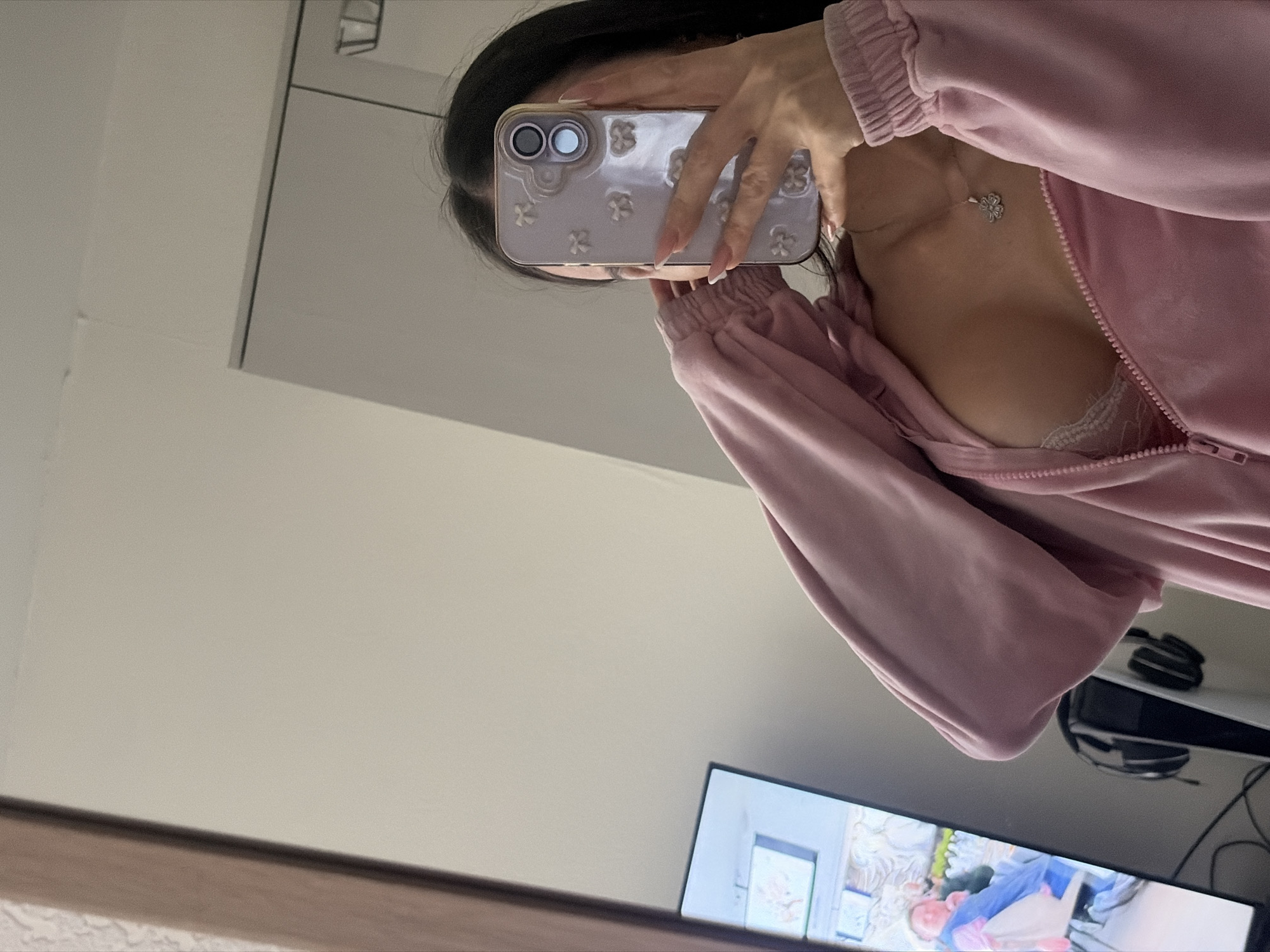 A female subject is depicted in a mirror selfie taken indoors. She has long dark hair and wears a pink zippered hoodie or tracksuit top, partially unzipped to reveal a white lace bra and a silver necklace with a four-leaf clover pendant. Her right hand, adorned with long, light-colored manicured nails, holds a light purple smartphone with a case featuring white bow-shaped embellishments, obscuring her face.
The environment, visible in the mirror's reflection, consists of a light-colored room. To the left, a flat-screen television displays a scene of a person with light hair wearing a blue smock, possibly in a salon setting, with a white bag nearby. Below the television, a white PlayStation 5 console rests, with black over-ear headphones positioned on it. Cables are visible on the floor near the console. A wooden mirror frame is partially visible on the far left, and part of a light-colored wardrobe or cabinet is seen on the far right. The specific location within Saint Petersburg, Russia, cannot be determined from the visible elements of this indoor scene.