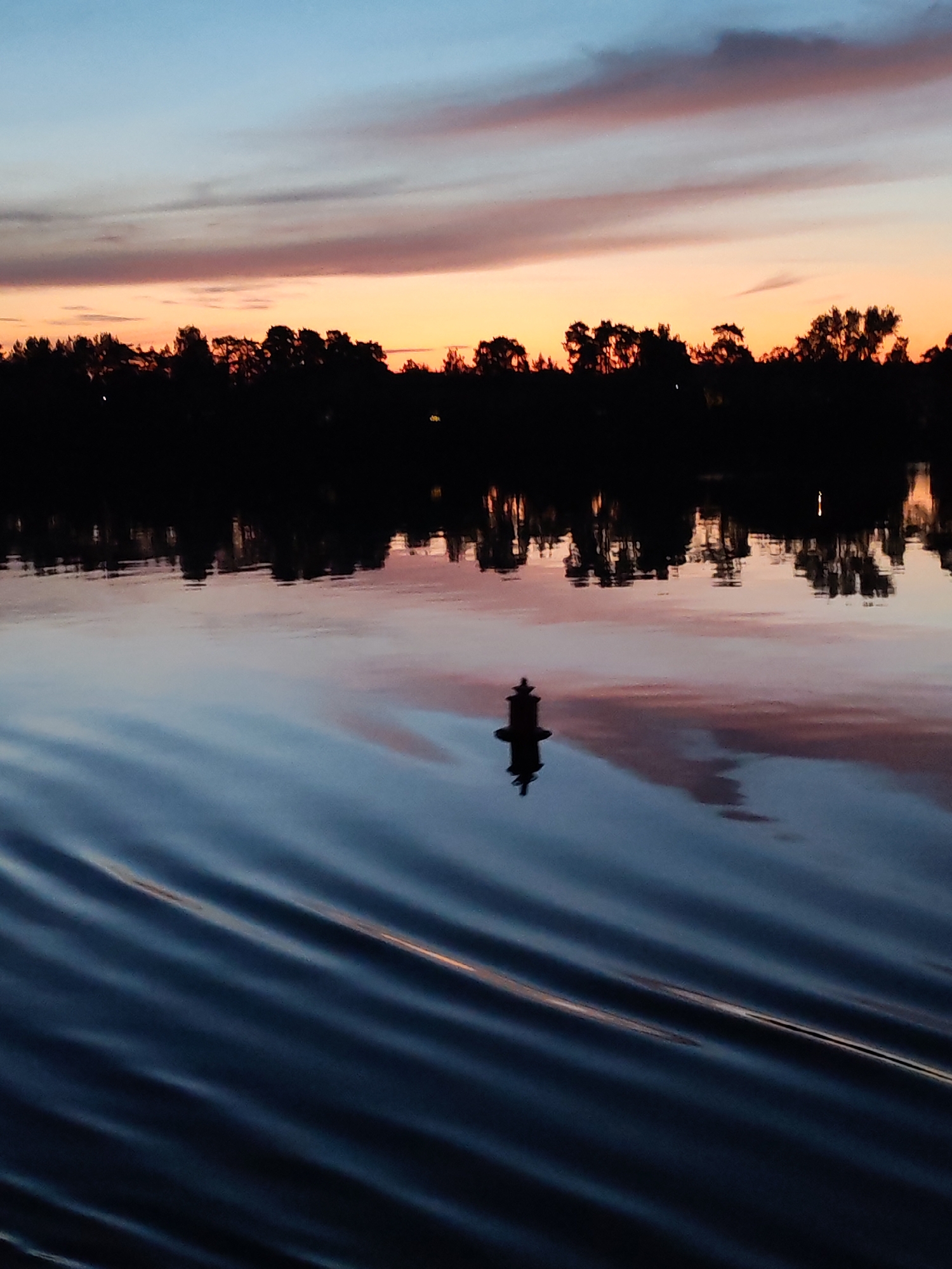 This is a vertical, medium-shot photograph taken at dusk or dawn of a serene body of water. The sky displays a gradient of colors, transitioning from a pale blue at the top to shades of pink and orange nearer to the horizon. Silhouetted against the sky is a dense line of trees, their forms dark and indistinct. The water's surface reflects the sky and the treeline, creating a mirror image. In the foreground, the water is rippled, showing texture and movement. A single, dark, buoy-like object, with a distinct shape, is reflected in the water, positioned off-center. The overall atmosphere is tranquil and peaceful.