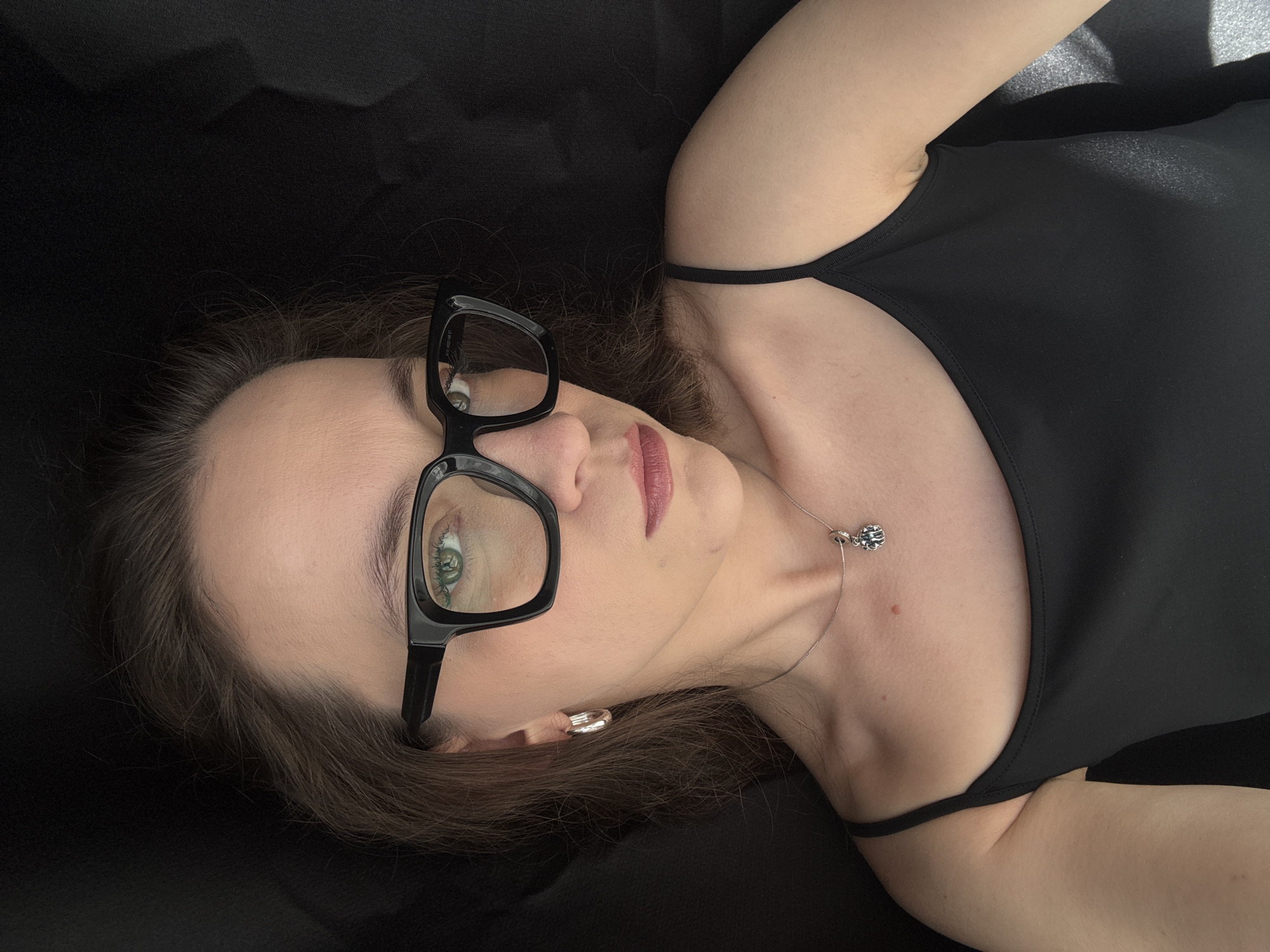 A woman with brown hair and green eyes, wearing black-rimmed glasses, a black tank top, and a silver necklace, is lying down on a black surface. Her head is tilted upwards, and she is looking towards the camera. The lighting suggests it is daytime, possibly indoors with artificial light or outdoors with diffused sunlight. Her facial expression is neutral. There is no visible text in the image. The setting appears to be a simple, dark background.