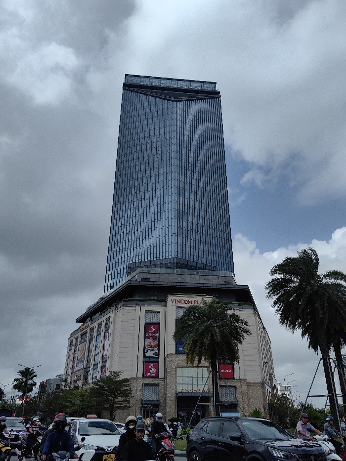 The image captures an urban street scene in Thuận Hóa Ward, Vietnam, featuring the Vincom Plaza building under a partially cloudy sky. The central subject is a tall, modern skyscraper with a dark blue, reflective glass facade, which rises above a wider, multi-story base structure. This base is constructed from light-colored stone or concrete, displaying "VINCOM PLAZA" signage prominently, along with various commercial advertisements, notably a "Jollibee" banner on one of its lower levels.
In the foreground, a busy city street is observed with a moderate volume of traffic. Numerous motorbikes are present, carrying riders who are predominantly wearing helmets; some also have face masks. A white sedan, potentially a taxi given its roof-mounted light, and a dark-colored SUV (identifiable as a black Peugeot) are also visible among the motorbikes. The traffic appears to be in motion or stopped at an intersection. Several large palm trees are interspersed throughout the scene, particularly on the right side and near the base of the Vincom Plaza building. The sky overhead presents a combination of dense grey clouds and visible patches of clear blue sky.