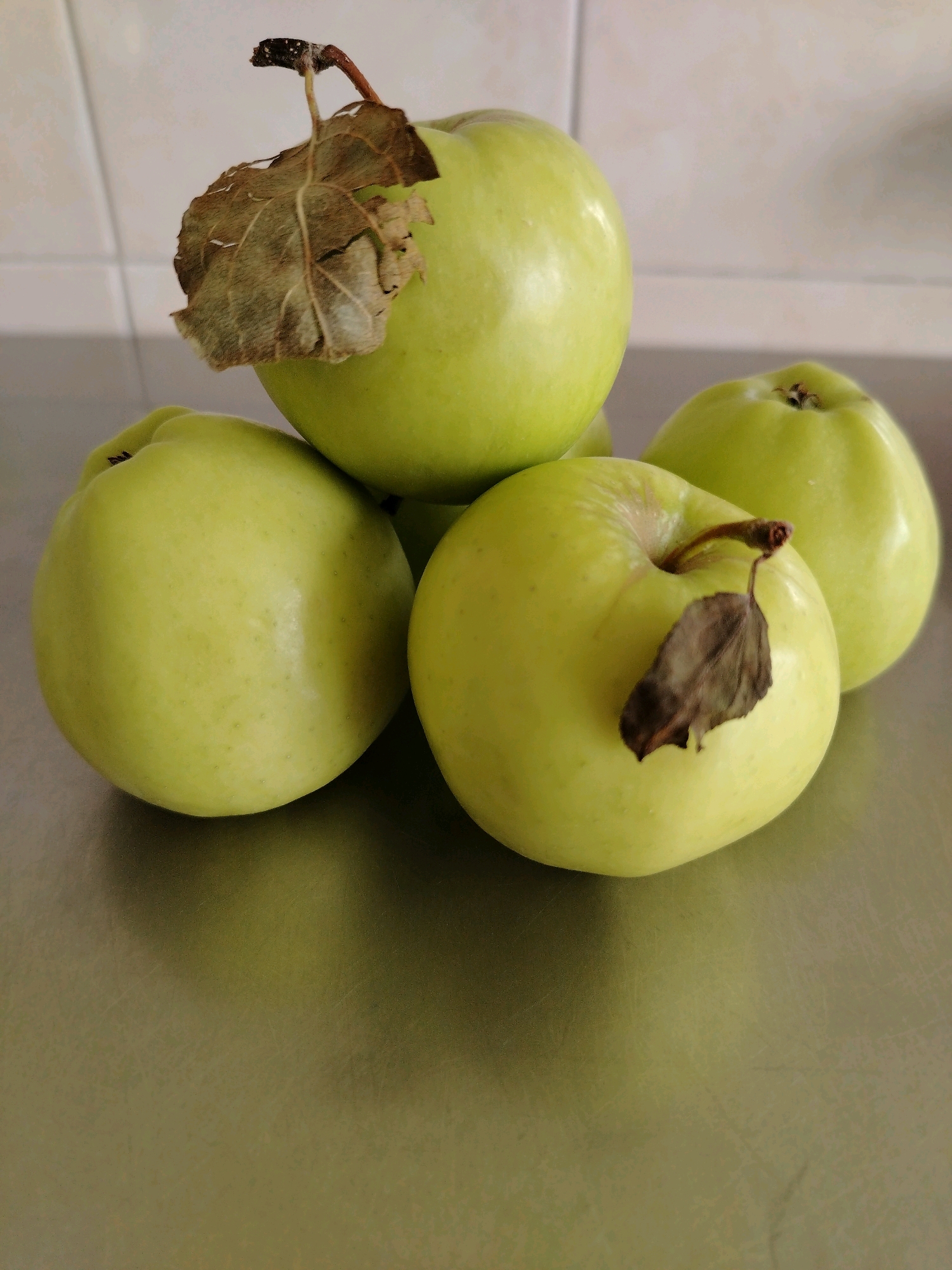 A still life image features four green apples arranged on a metallic surface, likely a countertop. Two apples are in the foreground, with one slightly in front and to the left of the other, and two more apples are positioned behind them. The apples appear fresh, with one in the front left and another in the back right having dry, brown leaves attached to their stems. The background consists of a tiled wall. There are no people or animals present. The scene appears to be indoors.