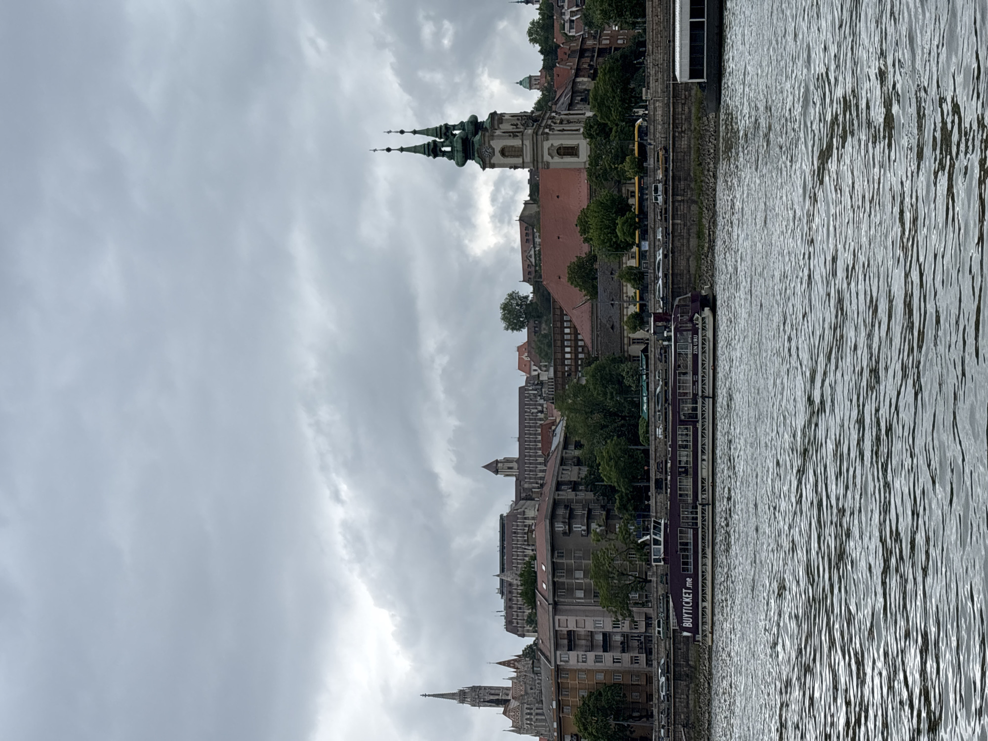 The image shows the cityscape of Budapest, Hungary, taken from a perspective on the Danube River. Prominently featured are the buildings along the riverbank, including various residential and commercial structures. Several church spires can be seen, including the Matthias Church, which has characteristic green, ornate spires. The sky is overcast and filled with clouds. The Danube River is in the foreground, reflecting light and showing water movement. A tourist boat is visible docked along the riverbank, with text reading "BUYTICKET.ME" displayed on the side.