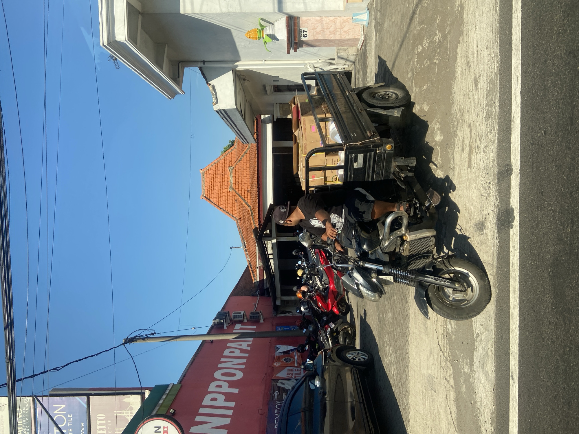 The image depicts a street scene in Ngrame, Indonesia, under a clear blue sky. In the foreground, a man wearing a black t-shirt, black shorts, and a black baseball cap is riding a black three-wheeled vehicle. The vehicle has a platform in the back loaded with cardboard boxes. The man is looking forward, and his left hand is on the handlebars. To the left of the vehicle, a dark-colored car is parked. Behind the car and the three-wheeled vehicle, several motorcycles are parked. A large red building with white lettering that reads "NIPPON PAINT" is visible on the left side of the image. The building has several air conditioning units mounted on its exterior. Further back, traditional Indonesian roofs with orange tiles are visible. To the right, a white building with a decorative pillar displaying the number "67" and an ornamental yellow object resembling corn on the cob is present. The ground is paved with concrete, and white lines indicating a crosswalk are visible in the lower portion of the image. Overhead power lines are also a prominent feature of the scene.