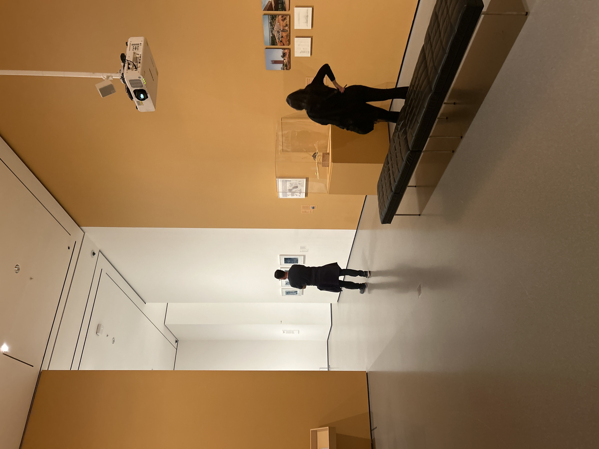 The image depicts an interior view of a museum, likely located in New York City, United States of America, as indicated by the architectural style and exhibits. The museum space features tan-colored walls and polished floors. In the foreground, a woman in a black outfit leans against a black leather bench, looking at artwork displayed on the wall. Another person, also in dark clothing, stands further down the hall, examining artwork. A projector is mounted on the ceiling, casting light in the direction of the tan wall. Several framed art pieces are displayed on the wall near the seated woman, and more art is displayed in the hall near the other person. The museum space also contains glass display cases. The environment is well-lit and appears to be a quiet and orderly museum setting.