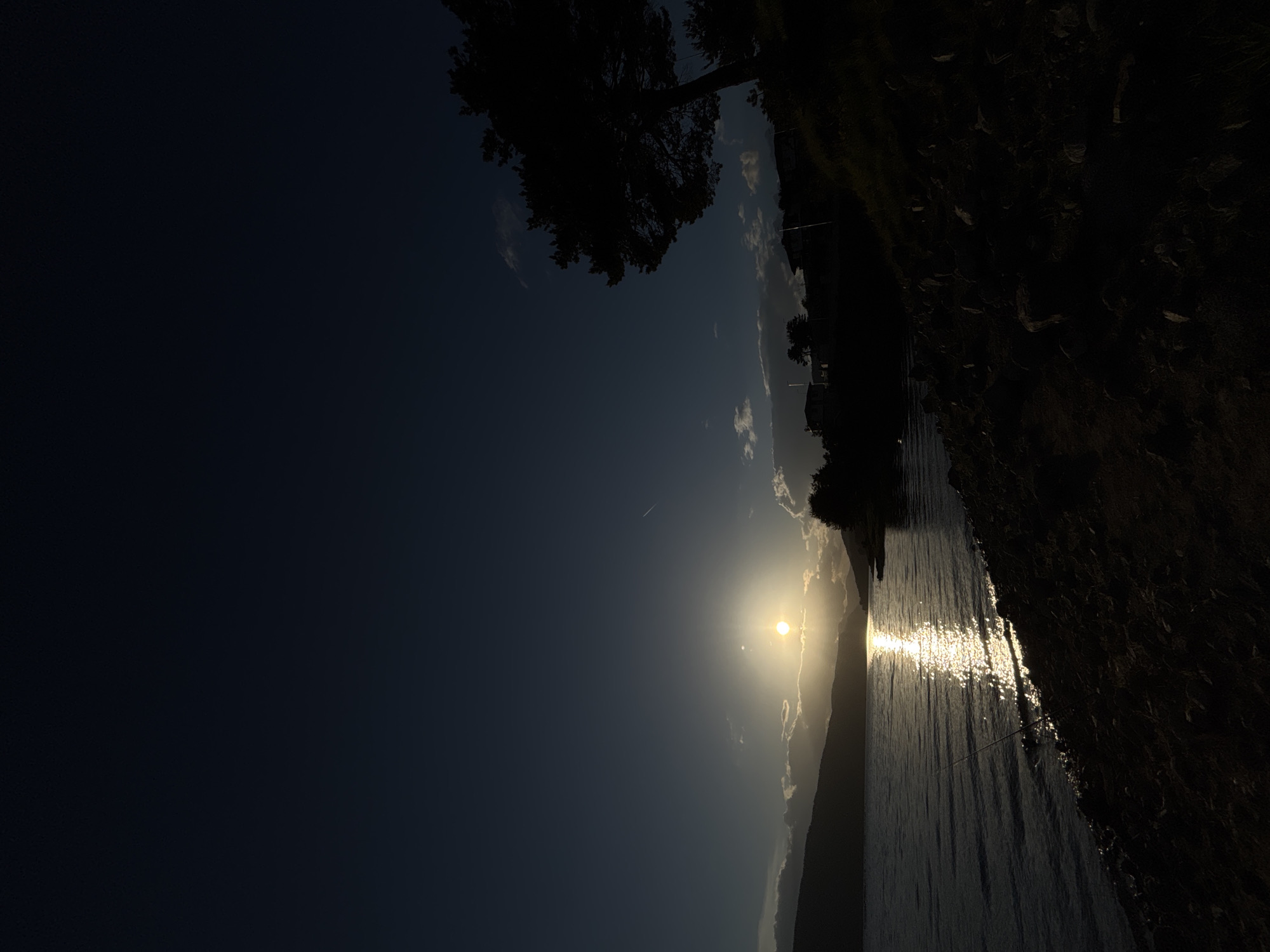 The image depicts a body of water reflecting sunlight, with a dark shoreline on the right side and silhouetted trees. The sky is dark with a bright sun obscured by clouds and landscape, creating a beam of light across the water. The location is unknown.