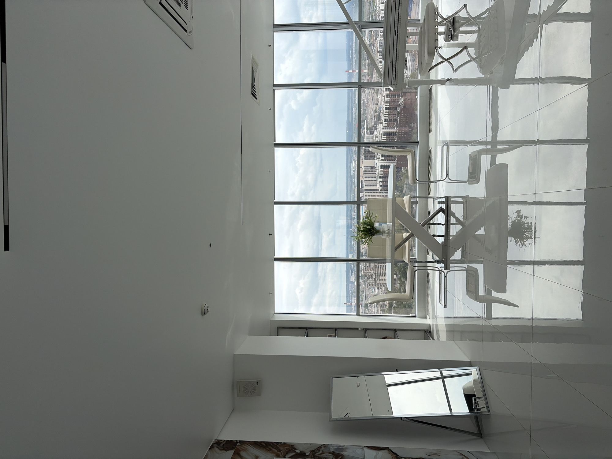 The image shows a well-lit, modern apartment with large windows overlooking a city skyline, likely Saint Petersburg, Russia, due to the architectural style and the presence of what appears to be the Neva River in the distance. The apartment features a white grand piano positioned to the right of a white dining table with chairs. A large, full-length mirror stands to the left, reflecting the interior and the city view. The flooring is highly polished and reflective, creating mirror images of the furniture and windows. The overall impression is one of spaciousness and contemporary elegance. The scene is devoid of people.