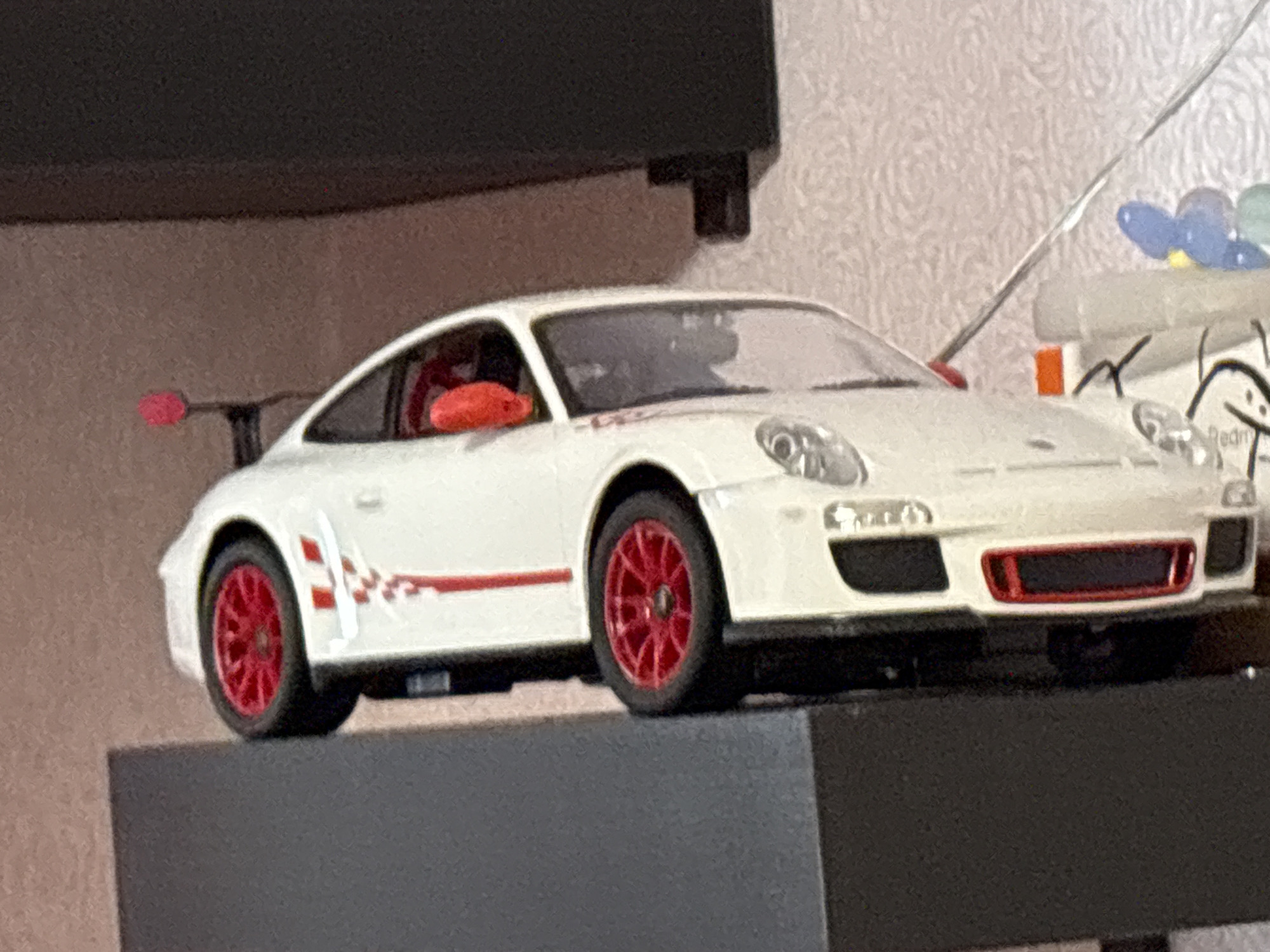 A white Porsche 911 GT3 RS model car with red wheels and accents is sitting on a gray surface. The car has a red racing stripe on the side and a red spoiler. The background is a textured wall and a dark object above the car. I can not identify the location in Прикамский, Russia as the photo lacks any information that could assist in this identification.