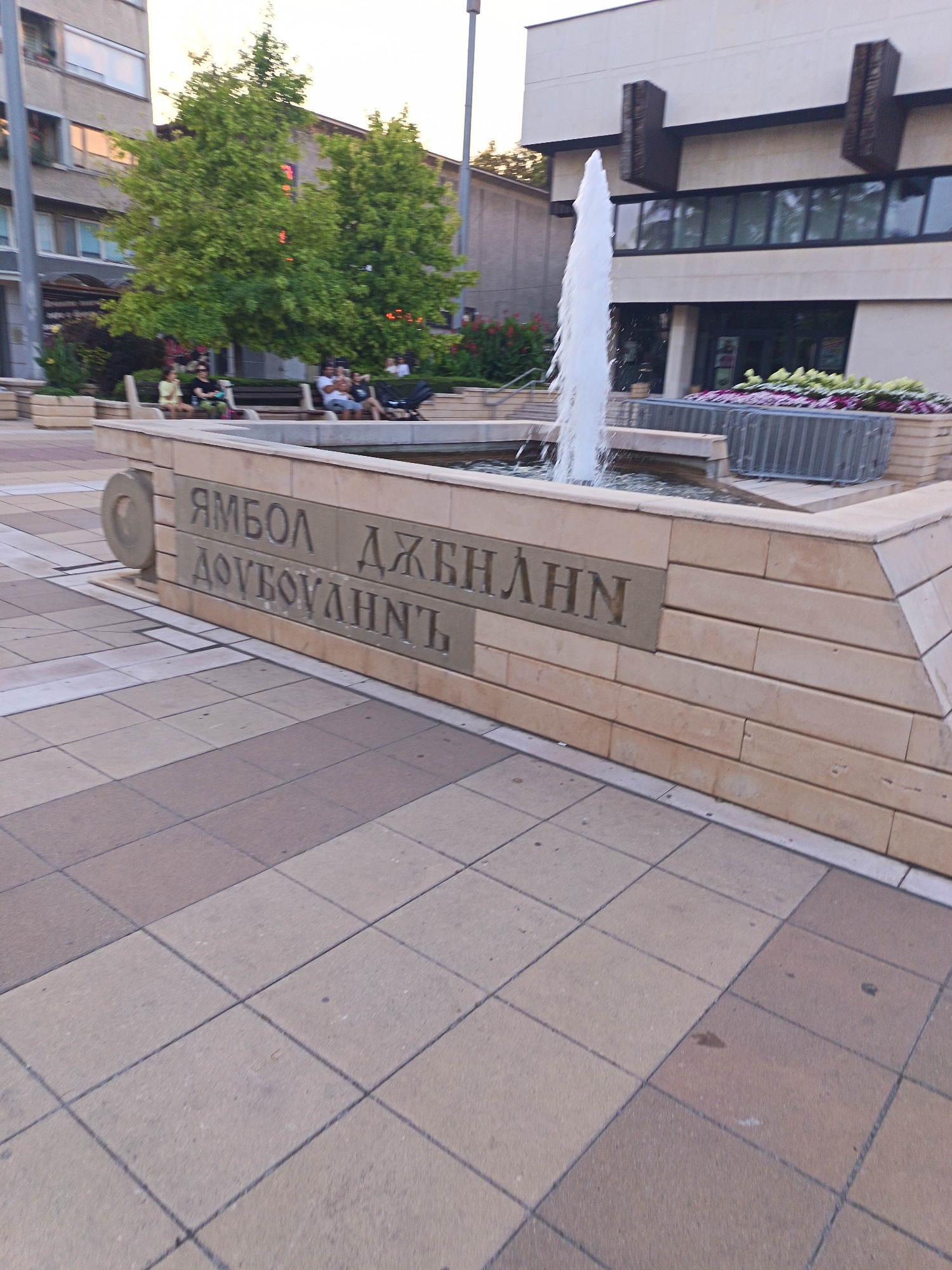 The image captures a view of a public space in Yambol, Bulgaria, featuring a fountain and inscribed stone blocks. The fountain, with water spraying upwards, is centrally located within a stone structure. The stone structure is composed of rectangular blocks, with some blocks featuring carved Cyrillic inscriptions. The inscription on the upper row reads "ЯМБОЛ ДЖЕНЛИНН", and the inscription on the lower row reads "ДОВГОВАЛНИ". In the background, a building with large windows and decorative elements is visible. Trees with green foliage are also present, along with people sitting on benches, suggesting a relaxed atmosphere. The ground is paved with square tiles in alternating shades of tan and brown. The scene appears to be in an outdoor urban setting.