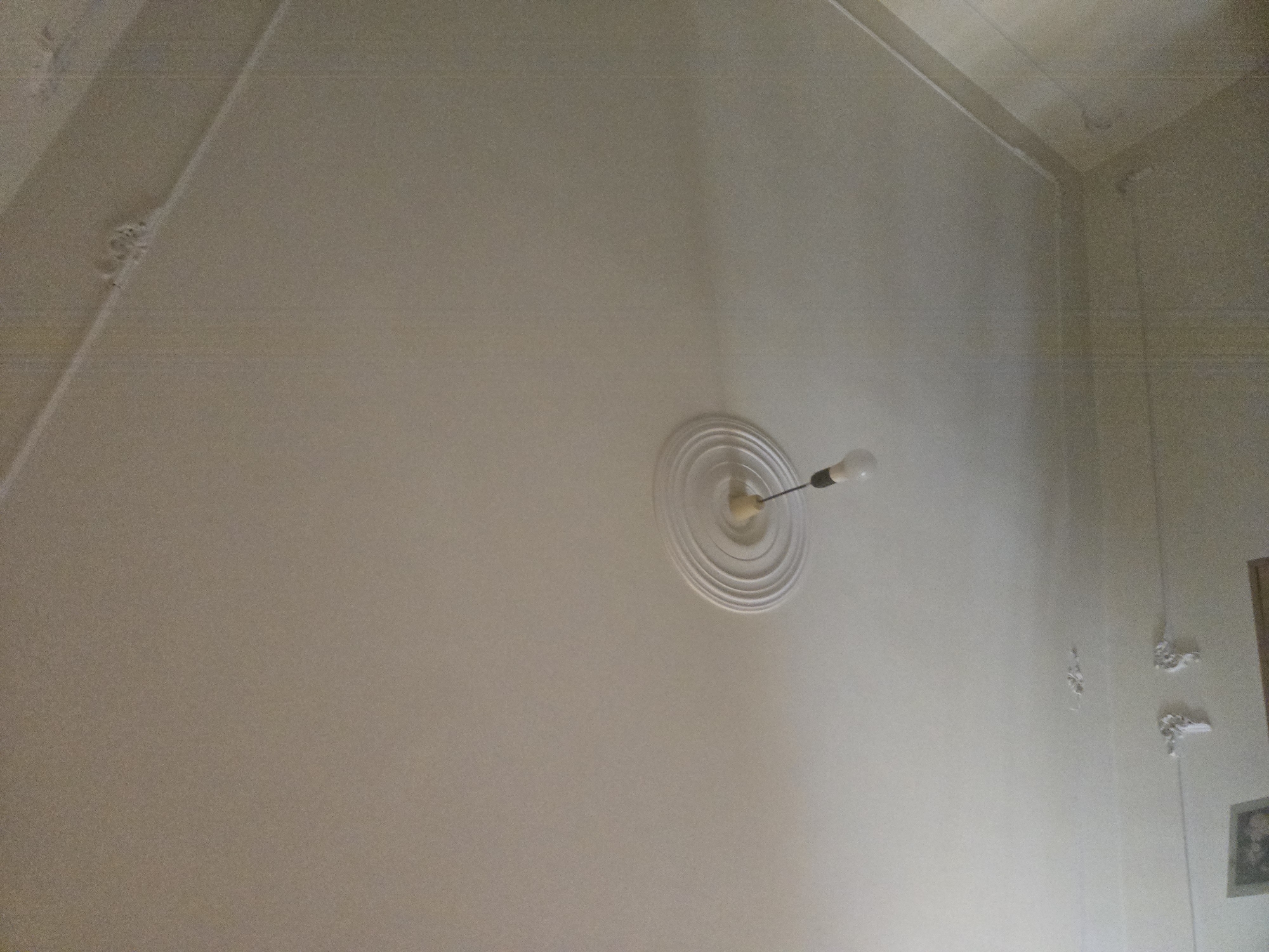 This image captures an indoor scene, primarily focusing on the ceiling and upper parts of the walls of a room in Yerevan, Armenia. The walls and ceiling are painted in a uniform, light off-white or cream color.
The most prominent object is a simple light fixture, consisting of a single bare light bulb hanging from the ceiling, which appears to be switched off. Surrounding the attachment point of the light fixture is a circular, multi-layered decorative ceiling medallion made of plaster.
The room features a complex ceiling structure, with sections that appear sloped or angled, particularly on the left side of the frame, suggesting it might be an attic room or a space under a pitched roof. In the upper left corner and along the upper right wall, there are areas of decorative plasterwork or potentially damaged plaster showing signs of wear or age. Thin lines, possibly electrical wires or tension wires, run along the intersections of the ceiling and walls.
On the far right edge of the image, a glimpse of a doorframe or the edge of a picture frame can be seen, with what appears to be a framed image partially visible below it. No people are present, and no specific activity is taking place. The general impression is of a modest, perhaps older, interior space. There is no visible text.