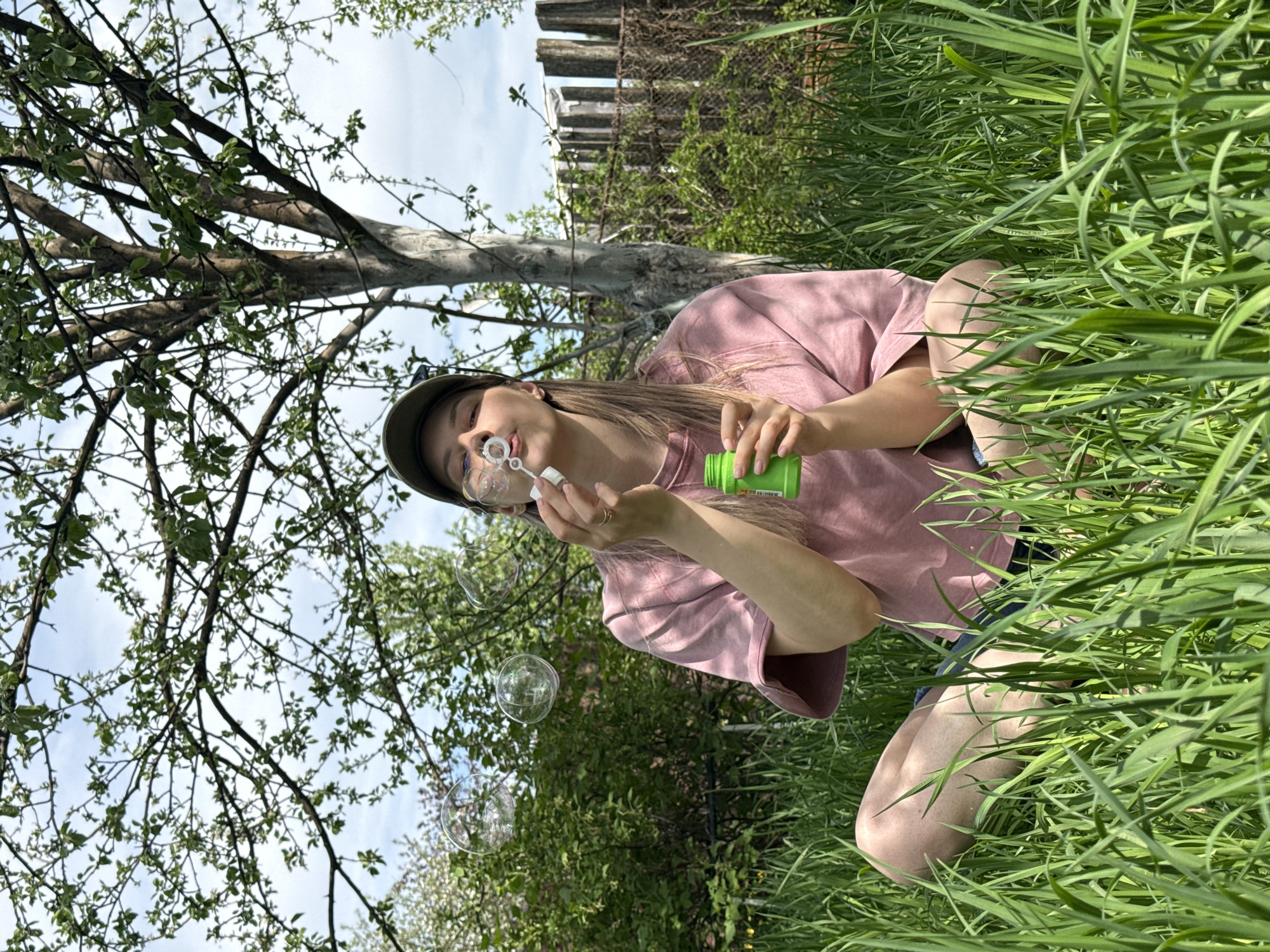 A woman is squatting in a grassy area, blowing bubbles. She is wearing a pink t-shirt, a dark-colored cap, and possibly shorts. She is holding a small green bottle in one hand and a bubble wand in the other. There are bubbles in the air around her. Tall green grass surrounds her, and behind her, there are trees, a fence, and a cloudy sky. This photograph was taken in the city of Chaikovsky, Russia.