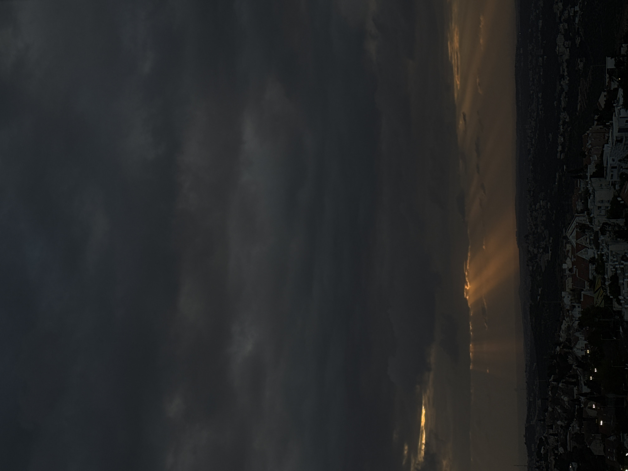 The image depicts a cityscape under a dark, cloudy sky with rays of sunlight breaking through. The location appears to be an urban area with numerous buildings, possibly houses or apartments, visible along the horizon. The sky is dominated by dark, heavy clouds, with a few streaks of golden light peeking through, creating a dramatic contrast. The light rays are casting shadows over parts of the city.