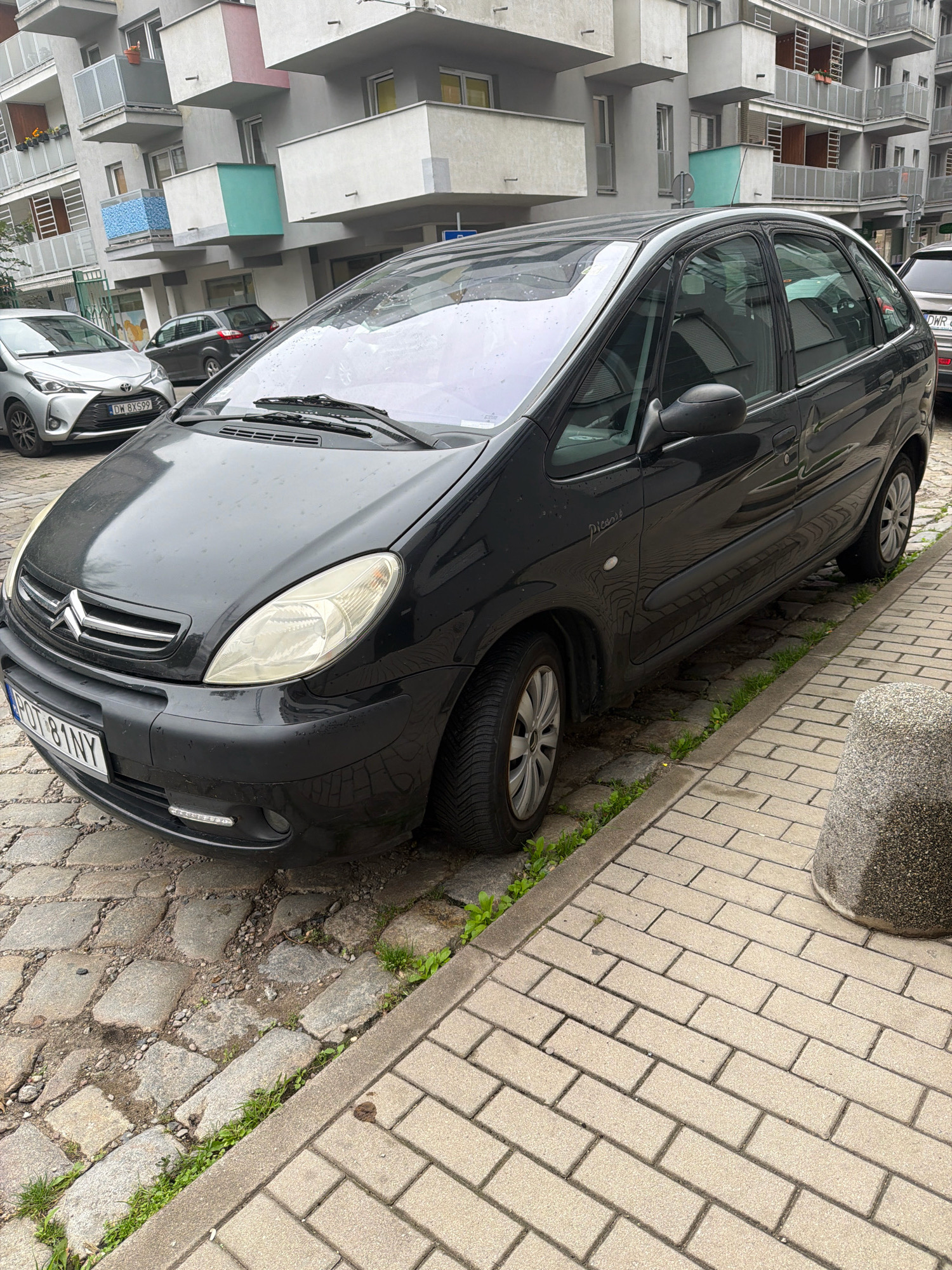 The image shows a black Citroën Picasso parked on a cobblestone street in front of a modern apartment building. The car is wet, suggesting recent rain. The building has multiple balconies and a light grey facade. A silver Toyota Yaris is parked behind the Citroën. The street is lined with a sidewalk made of beige bricks. In the foreground, a large, round, grey concrete bollard is visible. The scene appears to be in an urban residential area. The license plate of the Citroën indicates it is registered in Poland, likely Wrocław, given the characteristic yellow and black plates.