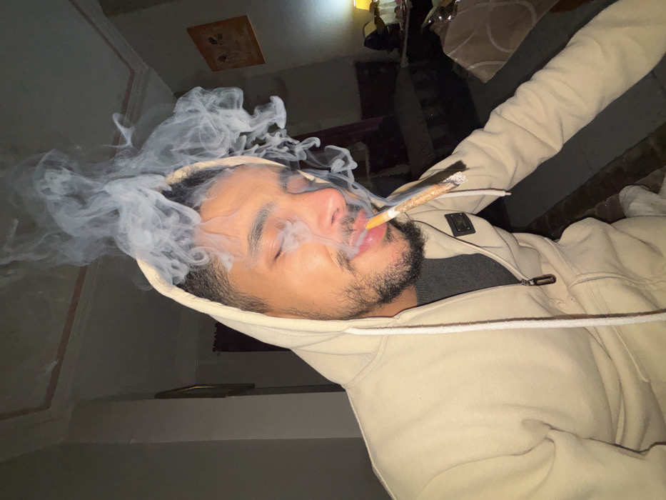An indoor photograph depicts a young adult male with a beard and closed eyes, wearing a light-colored hooded sweatshirt over a dark garment. A lit hand-rolled item, consistent with a cigarette or joint, is held between his lips, emitting a significant volume of white smoke that rises around and above his head. The background reveals a dimly lit residential interior in Tanger, Morocco, featuring light-colored walls with decorative molding, framed artwork, and dark upholstered furniture with cushions. A warm-toned lamp is visible on the right, providing ambient illumination. A small dark emblem is affixed to the right chest area of the subject's hoodie.