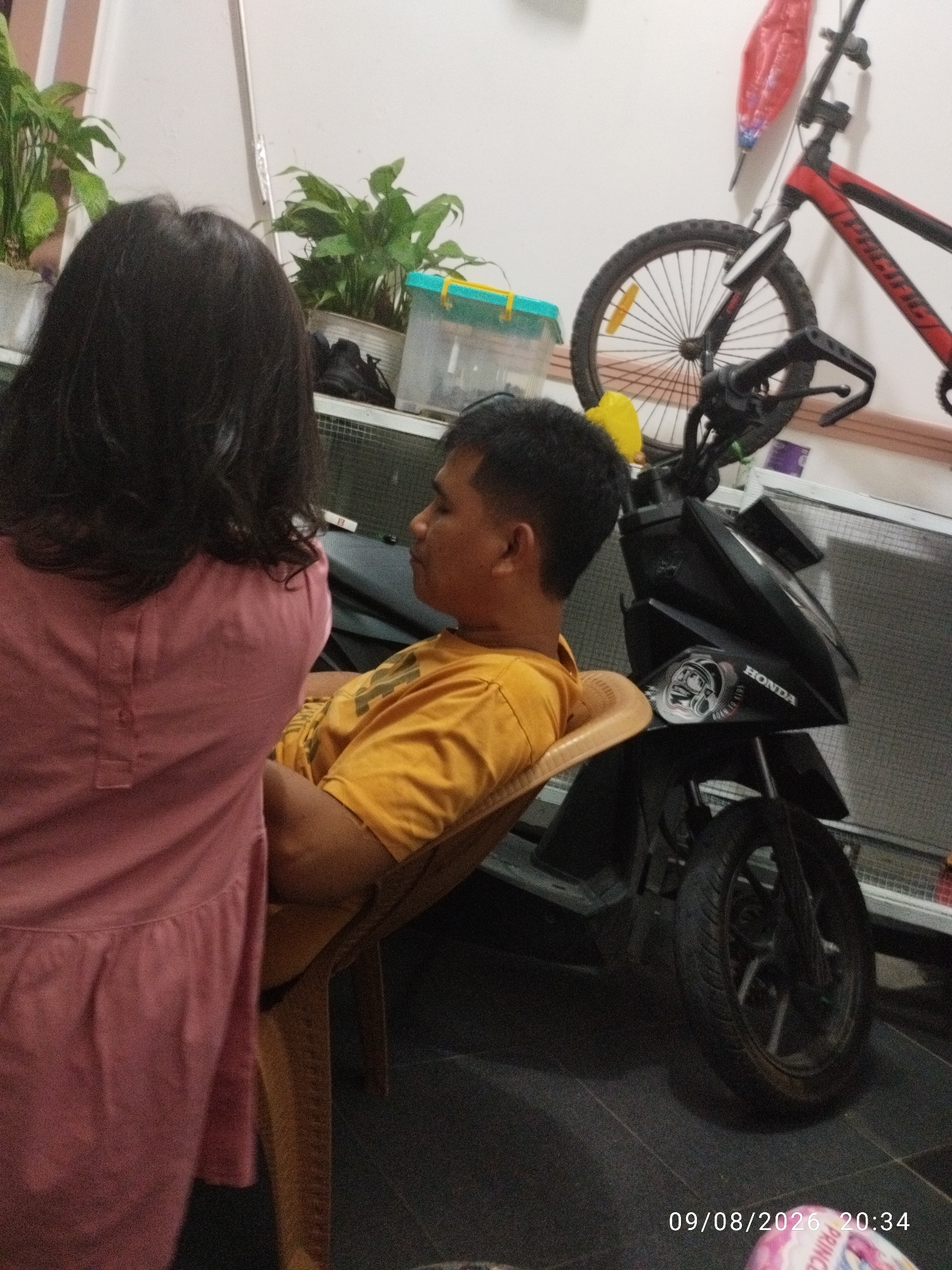 In Bandar Lampung, Indonesia, a young child, viewed from behind, stands near a man who is sitting in a chair. The child wears a pink dress, while the man is dressed in an orange t-shirt. They are positioned in a room with a dark tiled floor. To the right of the man is a black motorcycle, and behind it, a red and black bicycle leans against the wall. Green plants in pots are visible in the background, along with a blue and yellow plastic container. The scene is illuminated by indoor lighting. A timestamp indicates the image was taken on 09/08/2026 at 20:34.