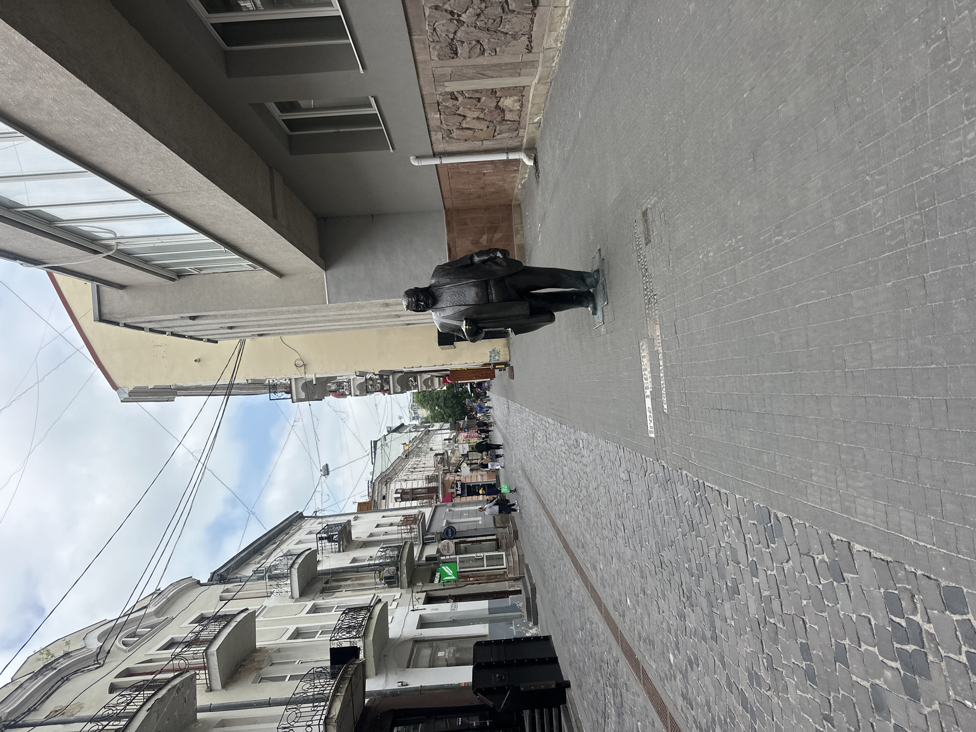 This image depicts a pedestrian street in Ternopil, Ukraine, featuring the Monument to Igor Hereta. The scene is observed from a slightly elevated, forward-looking perspective, capturing a street flanked by multi-story buildings under an overcast sky with visible patches of blue.
The street surface is bifurcated: the left and central sections are composed of dark gray cobblestones, with a narrow metal drainage grate running longitudinally down the middle. The right side features a smooth gray paved sidewalk, marked with a grid pattern.
On the right sidewalk, in the mid-foreground, stands a dark-colored statue of a robust male figure, identified as Igor Hereta by the inscription "Ігор Герета 1938-2002" visible on the pavement near its base. The statue portrays a man in a suit jacket over an embroidered shirt, holding a rectangular object (possibly a briefcase or book) in his left hand, with his right hand in his pocket. Behind the statue, a modern, multi-story building with a flat gray facade presents large rectangular windows. Its lower section is faced with reddish-brown, rough-hewn stone or concrete blocks adorned with carved relief patterns. A white vertical downspout is affixed to this wall. A lighter yellow building is partially visible further back on the right.
The left side of the street is lined with a series of 3-4 story buildings exhibiting classical architectural elements. These structures have light-colored stucco facades, multiple windows, and decorative black wrought-iron balconies. Their ground floors host commercial establishments, indicated by various signs, some in Cyrillic (e.g., a green logo, "ІLÉVEL").
Approximately 7 to 10 individuals are visible walking along the street in the mid-ground and background, generally moving away from the camera's viewpoint. They are casually dressed; one person in the mid-ground carries a green bag. Above the street, numerous black electrical or communication wires are strung across the thoroughfare. The lighting is diffuse and even, characteristic of a cloudy daytime environment.