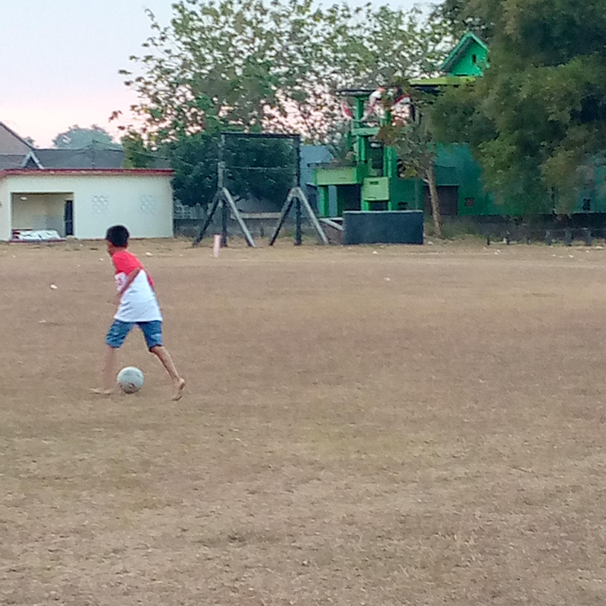 In Pacing, Indonesia, a young boy in a red and white t-shirt and blue shorts is playing soccer on a dirt field. The boy, barefoot, is in motion, kicking a light blue soccer ball. In the background, there is a low building with a white facade and a dark entrance, and behind that, a green structure, possibly a tower or a building with a small pitched roof. Trees with dense foliage are visible in the background, and some fencing and a net structure are also present. The sky is overcast and light. The ground is dry and covered with sparse, dry grass.