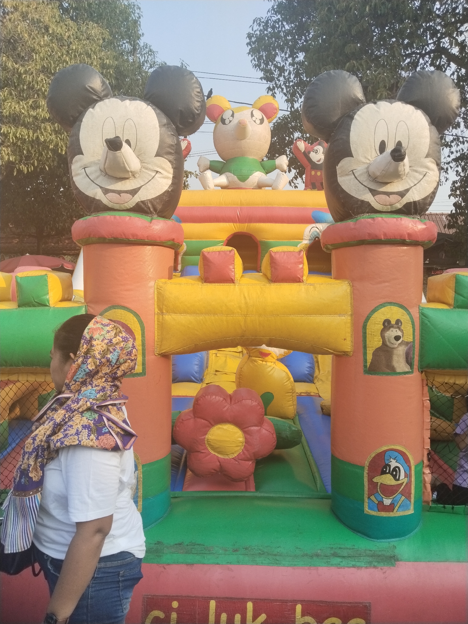 A bouncy castle, featuring characters like Mickey Mouse, a bear, and Donald Duck, is set up outdoors in Sragen, Indonesia. The inflatable structure is brightly colored with orange, yellow, green, and red sections. In the foreground, a woman wearing a white t-shirt, blue jeans, and a floral headscarf is standing with her side to the camera, looking towards the bouncy castle. The sky above is a pale blue, and trees are visible in the background. The ground in front of the bouncy castle is a mix of pink and green surfaces.