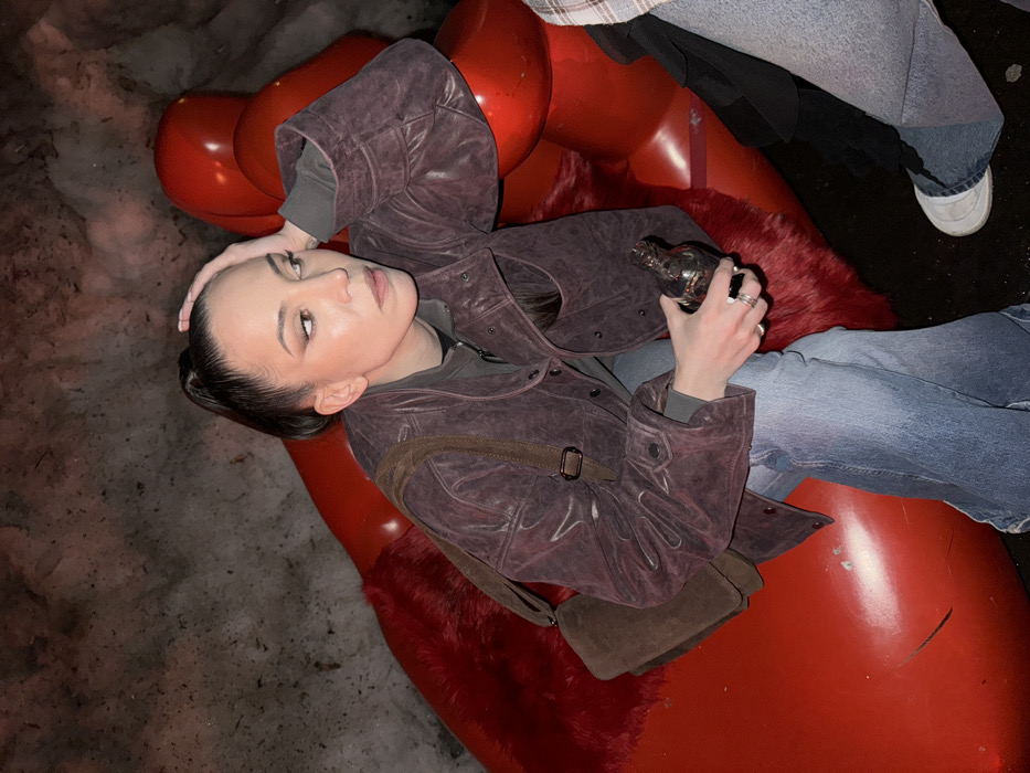 A woman with dark hair in a high ponytail is seated on a large, glossy red sculpture, identified as "The Red Hand" art installation, located in Zaryadye Park, Moscow, Russia. She is wearing a dark purple quilted jacket over a dark grey long-sleeved shirt, paired with light blue flared jeans. Her left hand is positioned on her head, while her right hand holds a small, uniquely shaped dark glass bottle. Multiple silver rings are visible on her left hand. A brown suede cross-body bag with a flap rests on her left side. She looks towards the viewer's left with a neutral, contemplative expression. A second individual's lower leg, clad in light blue jeans and a white sneaker, is partially visible on the far right, indicating they are also seated on the red sculpture next to the primary subject. The background consists of a snow-covered mound or wall, suggesting an outdoor, cold environment, with artificial lighting illuminating the scene.