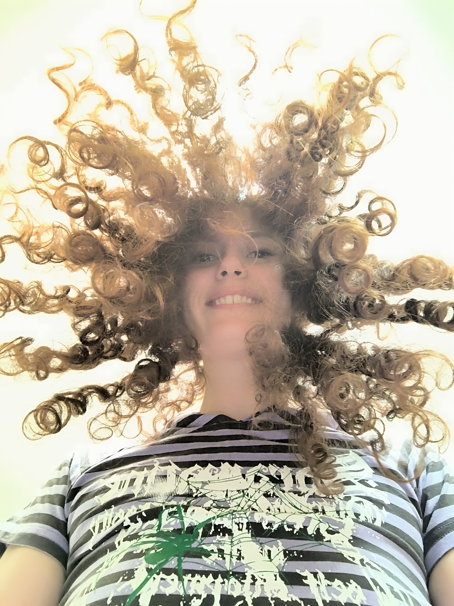 A low-angle shot depicts a person with a wide smile, their face partially obscured by a mass of curly, light brown hair that fans out around their head. The hair appears to be in ringlets, some catching the light and appearing lighter in color. The person is wearing a striped t-shirt in shades of purple, black, and white, with an abstract design in white and green on the front. The background is a solid, pale color, possibly white or off-white. There is no evidence to suggest that the location is in Saint Petersburg, Russia.