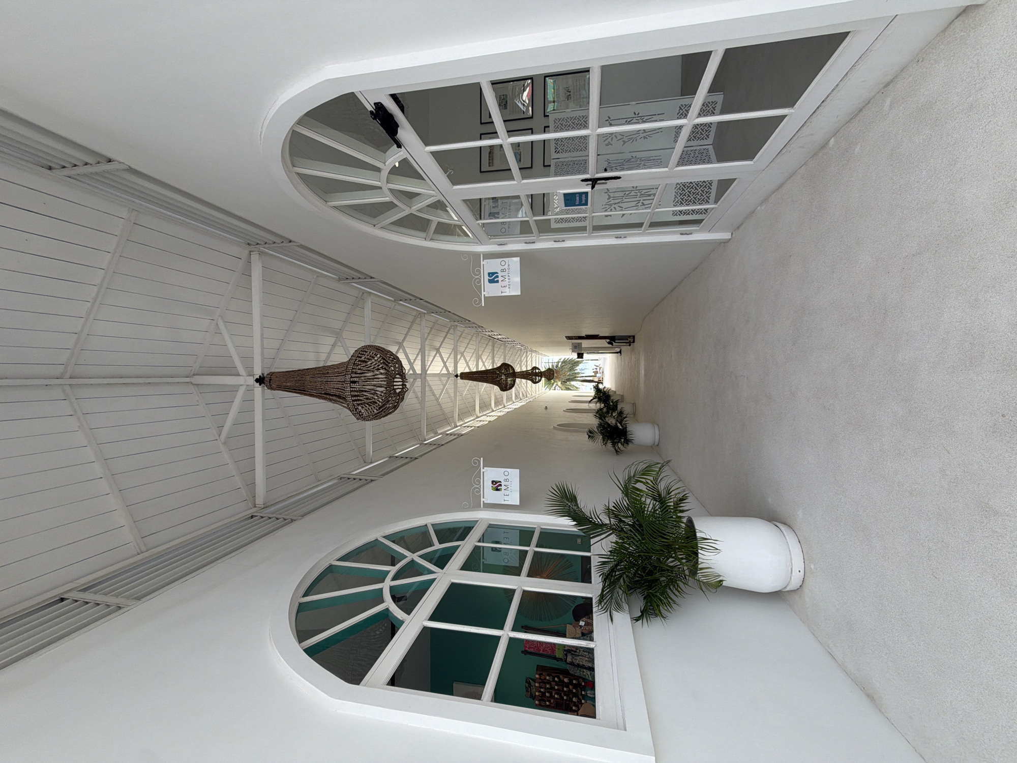 The image shows a long, white, outdoor hallway in Baan Bo Phut, Thailand. The hallway is covered by a white, slatted roof supported by white beams. Several large, beaded chandeliers hang from the ceiling. On either side of the hallway are arched doorways and windows, some of which appear to be entrances to shops or reception areas, as indicated by signage that reads "TEMBO RECEPTION". White potted plants with green foliage are placed along the hallway, adding a touch of nature to the minimalist architecture. The floor is a light-colored, textured surface. The overall impression is one of clean lines, bright light, and a serene, tropical ambiance.