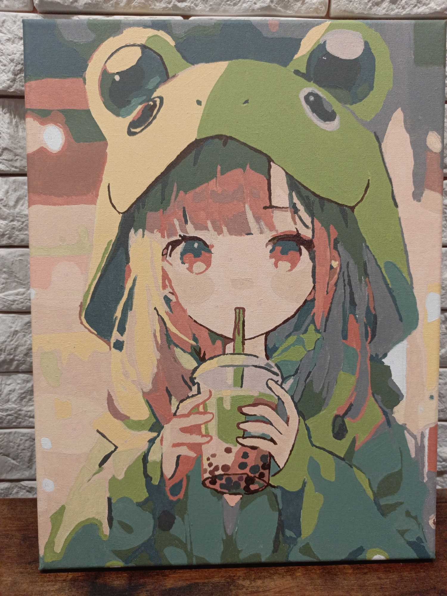 A rectangular painting rests on a wooden surface against a backdrop of a gray brick wall. The painting depicts an anime-style girl wearing a frog-themed hoodie, sipping from a cup of bubble tea. The hoodie is primarily green and yellow, with the frog eyes prominently featured. The girl has large eyes, fair skin, and hair that is colored in shades of pink, blonde, and gray. She holds the bubble tea with both hands, a straw positioned between her lips. The background of the painting has various muted tones including pink, peach, gray, and green. The painting has a "paint by numbers" aesthetic. There is no indication of the city of Chaikovsky, Russia in the image.