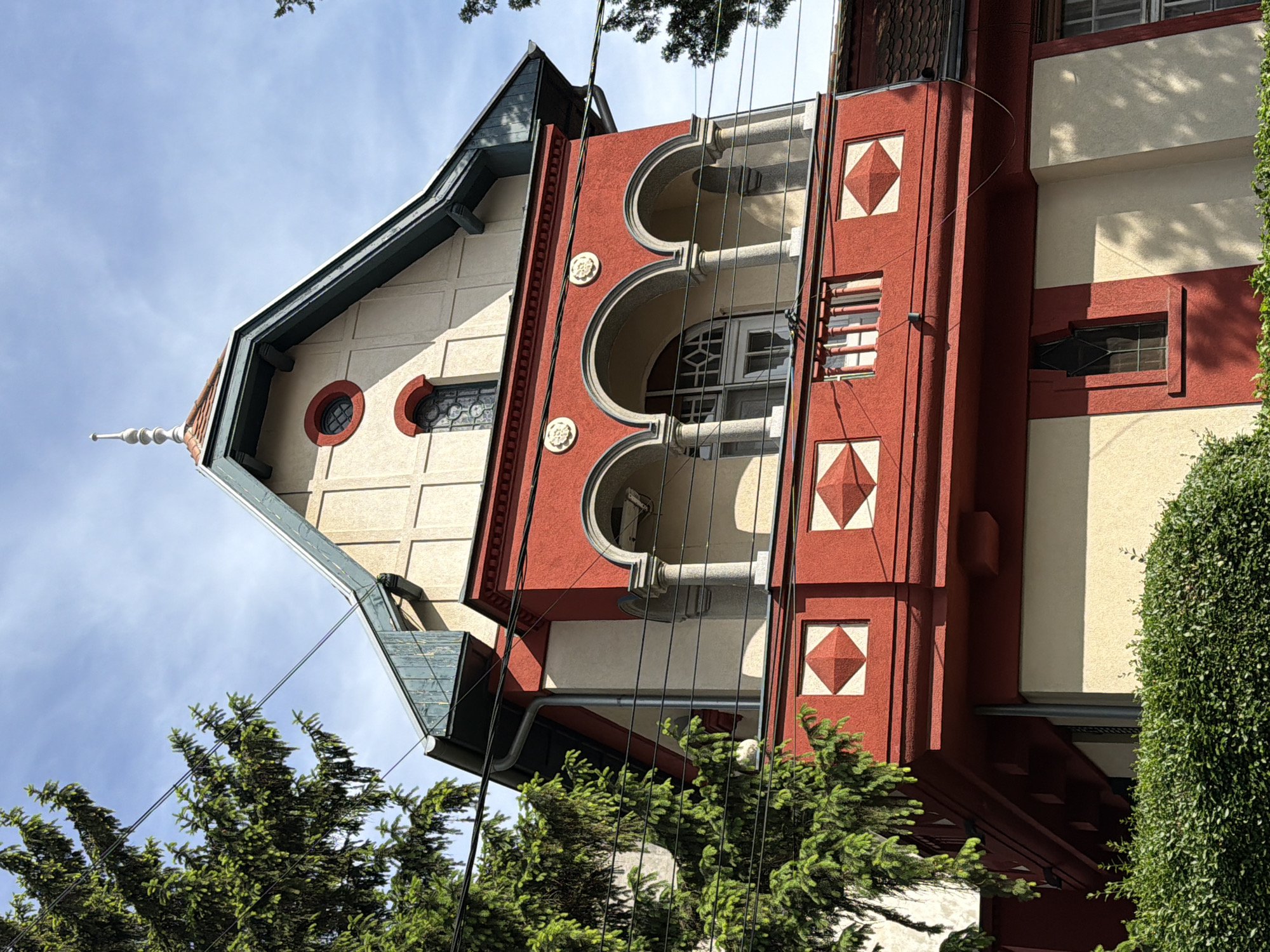A low-angle, medium shot depicts a section of an ornate building in Belgrade, Serbia. The building features cream-colored siding and red brick detailing. A pointed roof is visible at the upper left of the frame. The architecture features round window details, arched balconies with columns, and square diamond shapes as decoration. Power lines are visible crossing in front of the building. Tree branches are partially obscuring the base of the building. The sky is pale blue with light clouds.