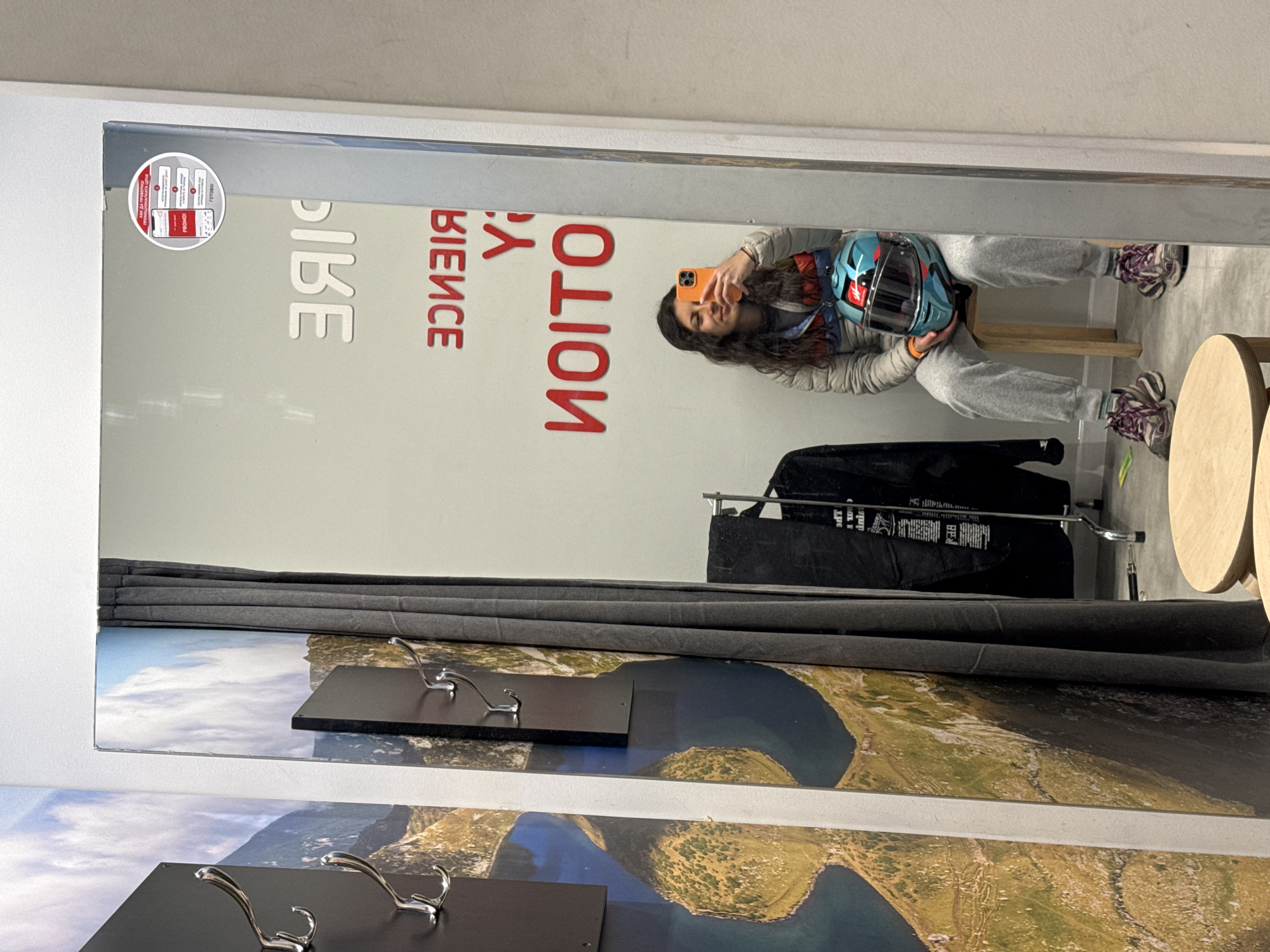 The image captures a person taking a mirror selfie in what appears to be a fitting room or a retail changing area, likely in Sofia, Bulgaria, given the visible Cyrillic text.
The primary subject is a person, possibly female, with long dark hair, wearing a light-colored puffer jacket, grey sweatpants, and purple and white athletic shoes. She is seated on a small wooden stool, holding a blue, red, and black motorcycle helmet with her left arm and an orange-cased smartphone in her right hand to take the photo. Her expression is a slight smile.
The mirror reflects the person and prominent text written on the wall behind them. The large red text, reversed in the mirror, spells out "МОТИОН" (MOTION) and "ЕКСПЕРИЕНС" (EXPERIENCE) when read correctly. In the top left corner of the mirror, a circular sticker is visible, displaying information in Bulgarian: "КАК ДА ВЗЕМЕШ ПРОМОЦИОНАЛНА ЦЕНА" (How to get a promotional price), with subsequent steps showing a phone screen with "PROMO!" and the final step saying "ГОТОВО!" (DONE!).
Below the mirror, the wall is decorated with a large panoramic image depicting an aerial view of a landscape featuring lakes and mountains. The foreground of this decorative panel includes two black rectangular fixtures with chrome coat hooks. To the right, partially visible, are sections of two round wooden tables or stools. A metal clothing rack with dark garments hangs behind the seated person. The floor is light-colored, suggesting concrete or tiled flooring.
The scene is indoors, with no specific time of day or weather discernible. The overall atmosphere appears casual and relaxed, indicative of a shopping or try-on experience.