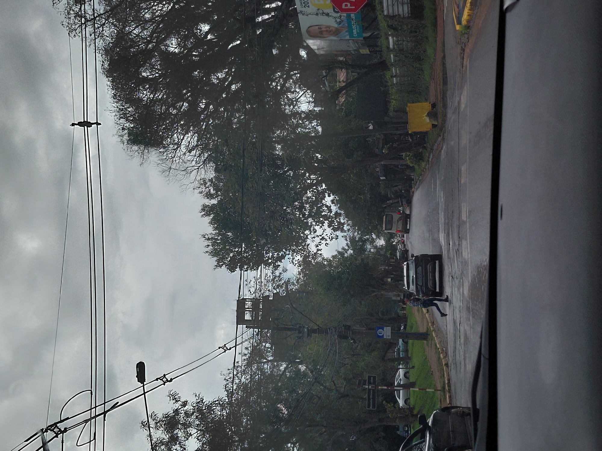 The image depicts a street scene in Encarnación, Paraguay, under an overcast sky. The view is from inside a vehicle, looking forward through the windshield and dashboard. A road with wet asphalt stretches into the distance, lined by trees on both sides. Power lines crisscross overhead.
In the foreground, a person is walking across a pedestrian crossing. They are wearing a checkered shirt and jeans. A dark-colored SUV is stopped near the pedestrian crossing, possibly yielding to the pedestrian. Several other vehicles, including a bus and cars, are visible further down the road.
On the right side of the road, a large billboard features a portrait of a man with the text "CARLOS GALEANO" and "CONCEJAL 2023". A red stop sign is also visible near the billboard. On the left side of the road, a street sign is partially visible. A blue sign with the letter "E" is posted on a utility pole. A utilitarian yellow structure is situated on the side of the road. The environment suggests an urban or suburban setting.