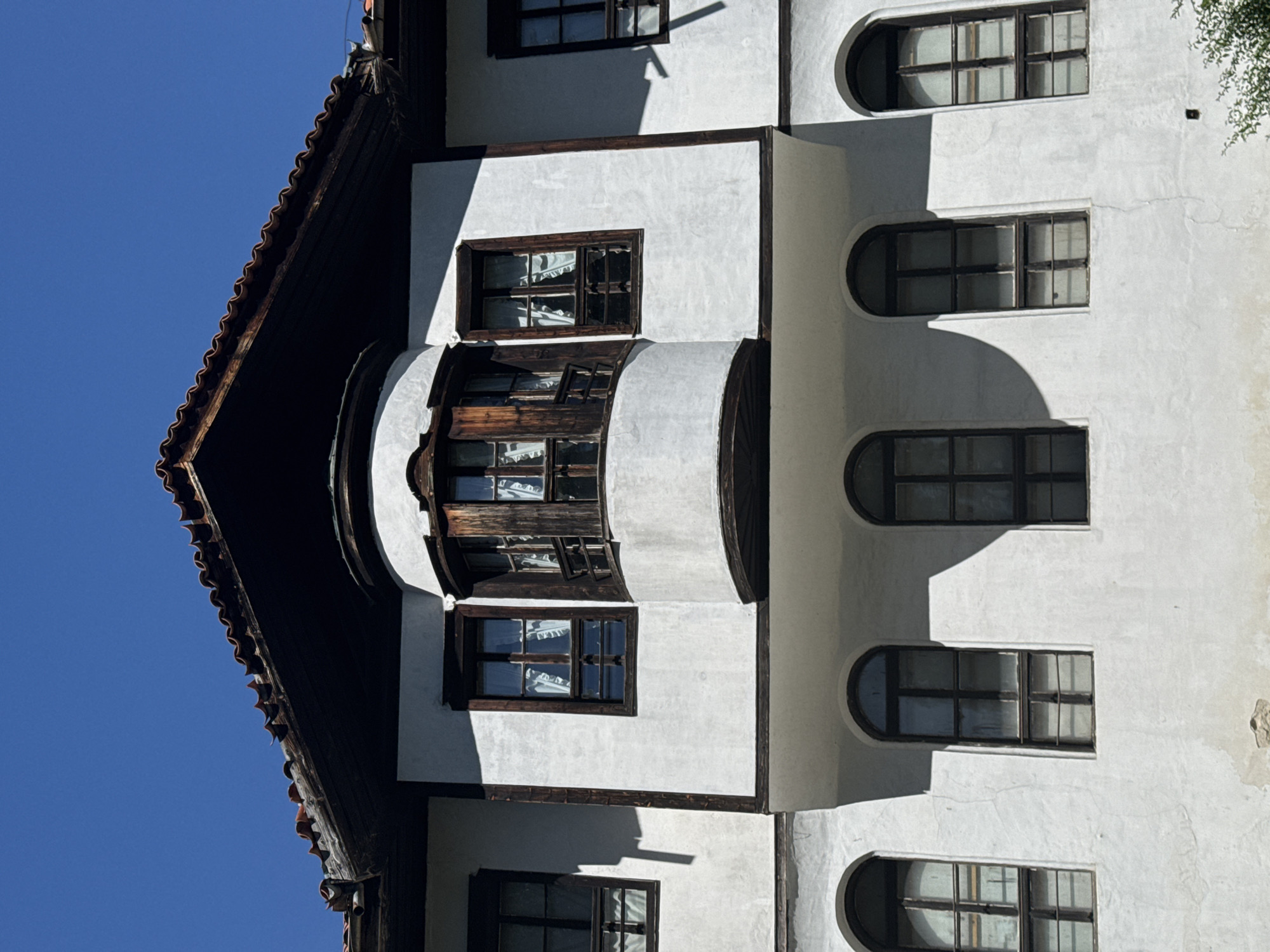 A white stucco building with dark wooden trim and window frames stands against a clear blue sky. The building features a multi-level facade with several windows. The uppermost level has a prominent, rounded bay window with wooden mullions and visible white curtains, flanked by two rectangular windows. A dark, overhanging roof with terracotta tiles caps the building. Below this, the second level displays three arched windows, each with dark frames and a grid of smaller panes, also covered by sheer white curtains. The wall surfaces are textured and show some signs of age. The sunlight casts distinct shadows on the building's facade.