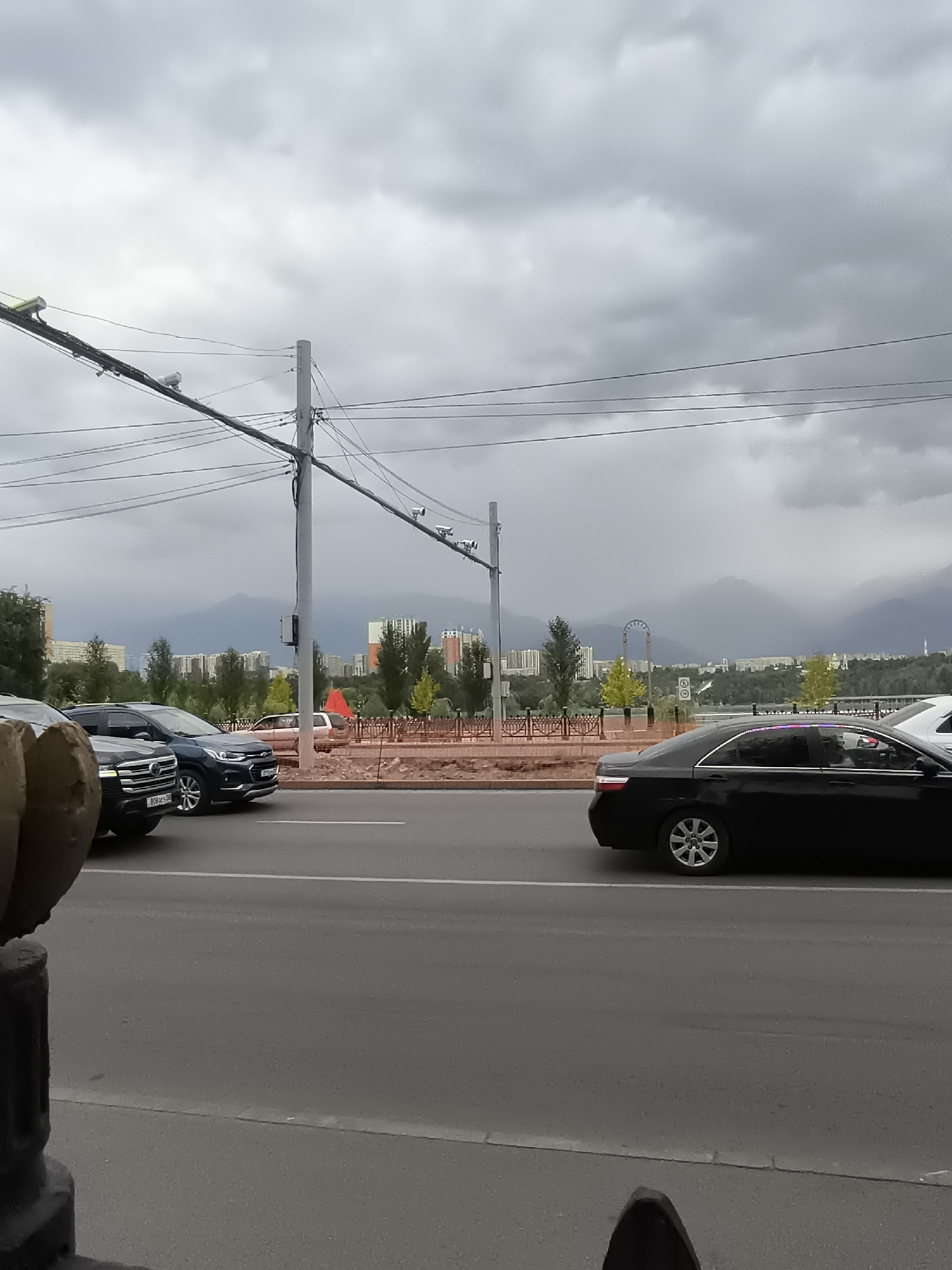 The image depicts a street scene in Almaty, Kazakhstan, under a heavily overcast sky. In the foreground, a dark-colored sedan is visible on the right side of the road, its wheels indicating movement. To the left, a line of cars is stopped at a traffic light, including a black SUV and a dark blue SUV. The road is paved, with a white lane marker.
Behind the road, a raised pedestrian walkway or park area is bordered by a decorative metal fence and a brick wall. Several trees and some potted plants are present in this area. In the background, a city skyline is visible, characterized by apartment buildings of various heights and architectural styles. Beyond the city, a range of mountains forms a dramatic backdrop, their peaks obscured by atmospheric haze or low-hanging clouds.
Several utility poles with an intricate network of wires stretch across the upper portion of the image. Security cameras are mounted on some of these poles. A bright red flag is planted near the pedestrian area. The overall impression is of a typical urban environment during a cloudy day.