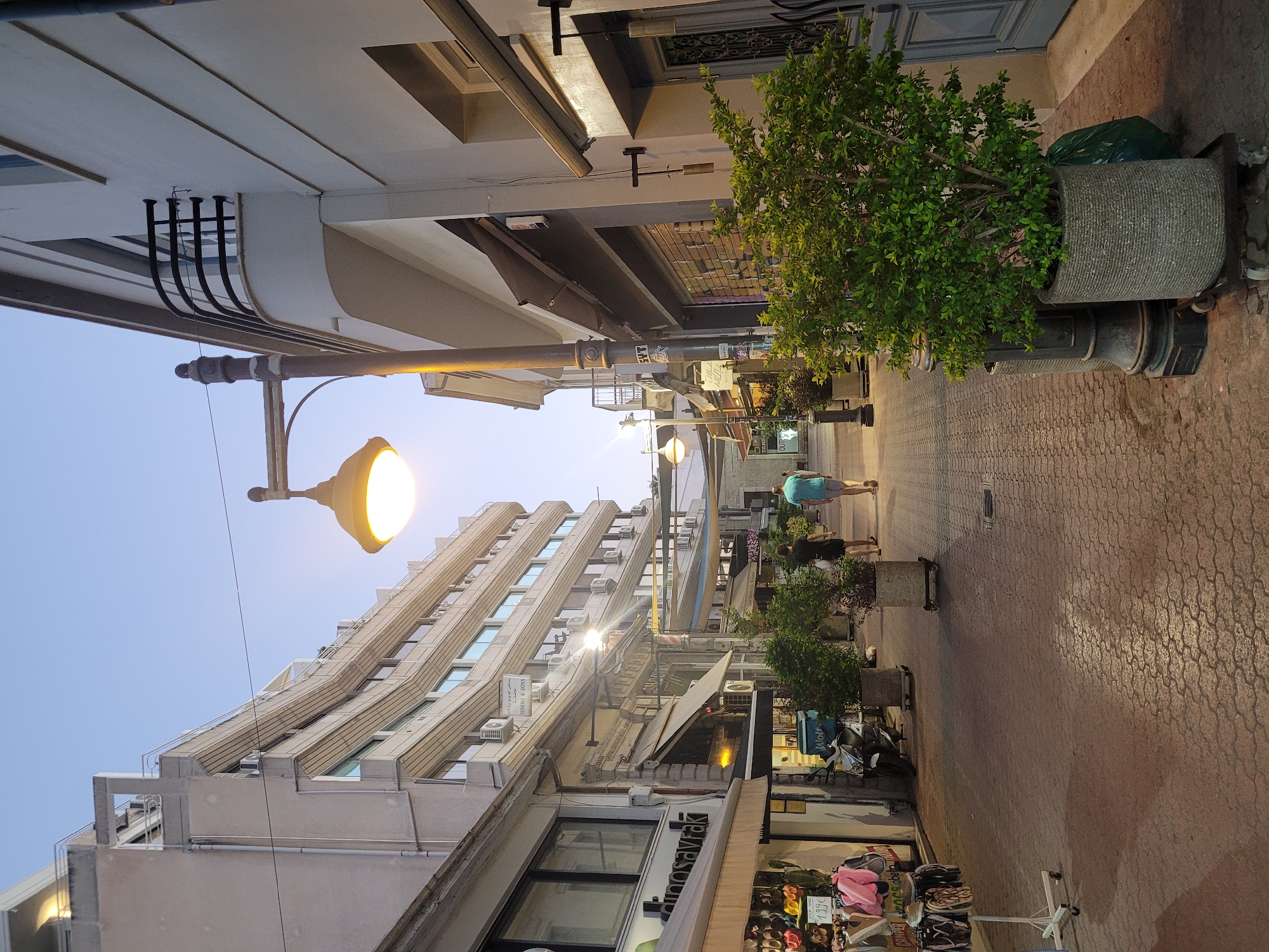 This is a photograph of a pedestrian street in Limassol, Cyprus, captured during twilight. The street is paved with interlocking bricks and lined with shops and buildings on both sides. A prominent street lamp with a glowing yellow light illuminates the scene.
On the left side of the street, there is a multi-story building with a curved facade, featuring numerous windows and air conditioning units. Below this building, shops display merchandise, including racks of footwear and clothing. A white display rack with flip-flops is visible. A scooter is parked in front of one of the shops.
On the right side of the street, a white building with a dark-colored door and a black wrought-iron balcony is present. Shops with awnings are also visible, some with their security shutters partially lowered. A large potted green bush stands in the foreground on the right.
Two individuals are walking away from the camera down the street. One person is wearing a dark t-shirt and shorts, and the other is wearing a light blue t-shirt and shorts. The sky above is a pale blue, indicating the transition from day to night. The overall impression is of a tranquil evening in a European city.