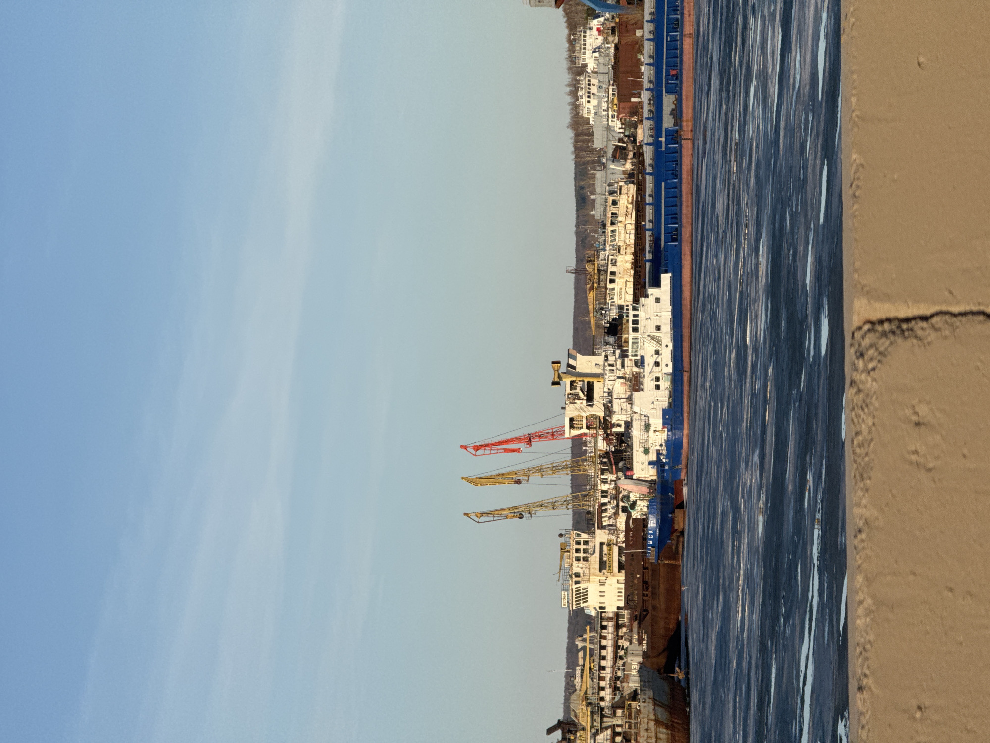 The image depicts a port scene in the city of Chaikovsky, Russia. Several ships and cranes are visible, docked along a body of water that appears partially frozen. The sky is clear and blue. A beige concrete structure is in the right foreground.