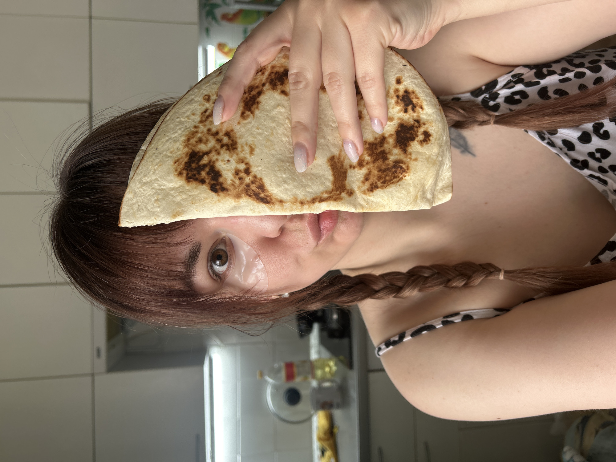 A female subject, appearing to be in a kitchen in Фоки, Russia, is depicted in a close-up, frontal portrait. She has brown hair with bangs and two visible braids. Her right eye is open, looking directly forward, with a translucent under-eye patch applied below it. The left side of her face is obscured by a folded, toasted flatbread, held by her right hand. Her lips, visible on the left, are slightly puckered. She wears a white top with black irregular spots. A dark tattoo is partially visible on her upper left chest/shoulder. Her right hand displays nails with a light, shimmery polish. The background reveals a kitchen environment with white cabinetry, a white tiled backsplash, a countertop, and a horizontal LED light strip. Objects on the countertop include a white kitchen appliance, a white mug, a clear plastic bottle with yellow liquid, and a black stovetop.