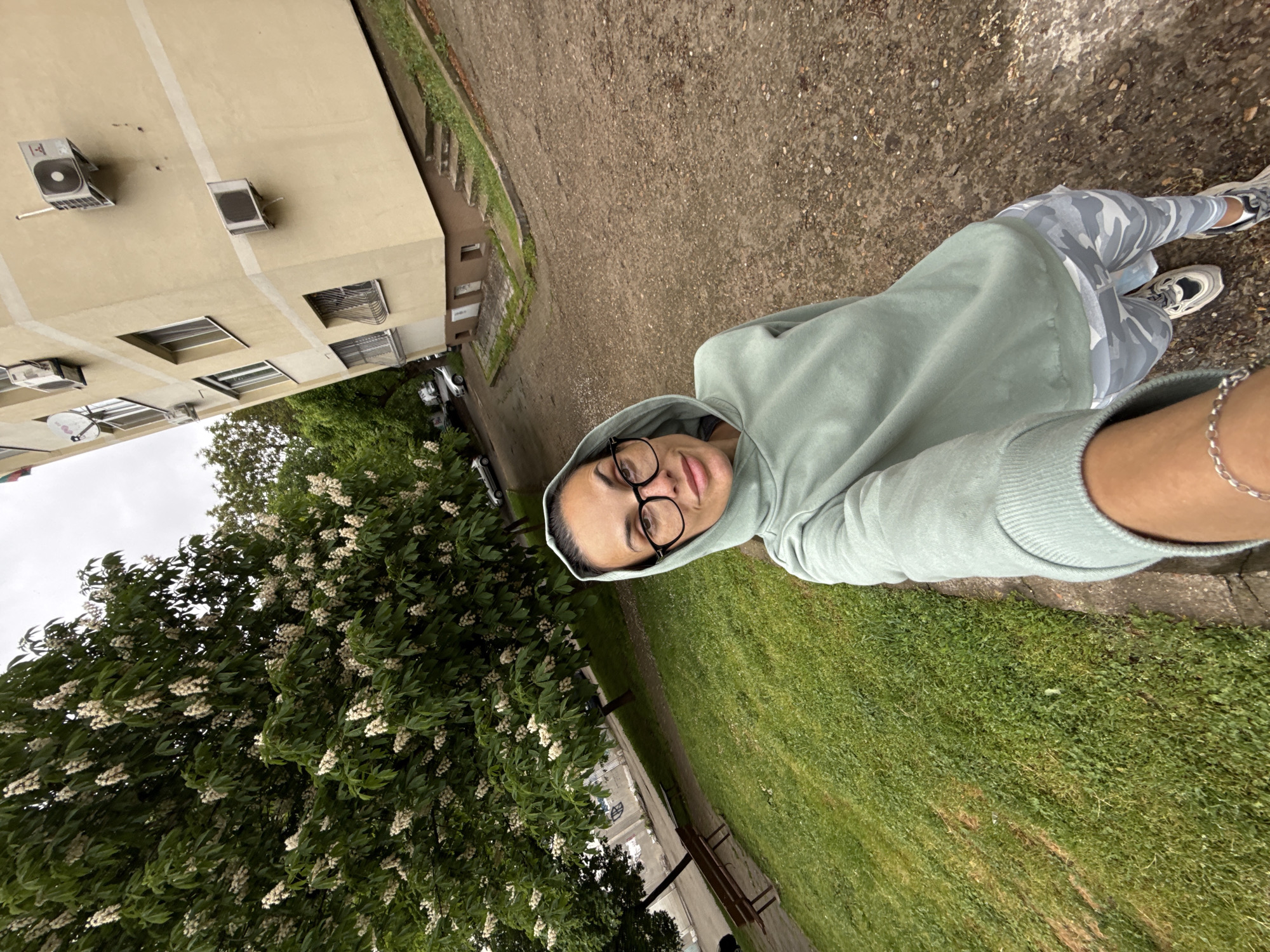 A selfie of a woman wearing a light green hoodie and glasses, standing outdoors. She is positioned on a paved surface near a grassy area and a large tree with white flowers. A light-colored building with multiple air conditioning units is visible in the background. The location appears to be a park or public space in an urban area of Pleven, Bulgaria.