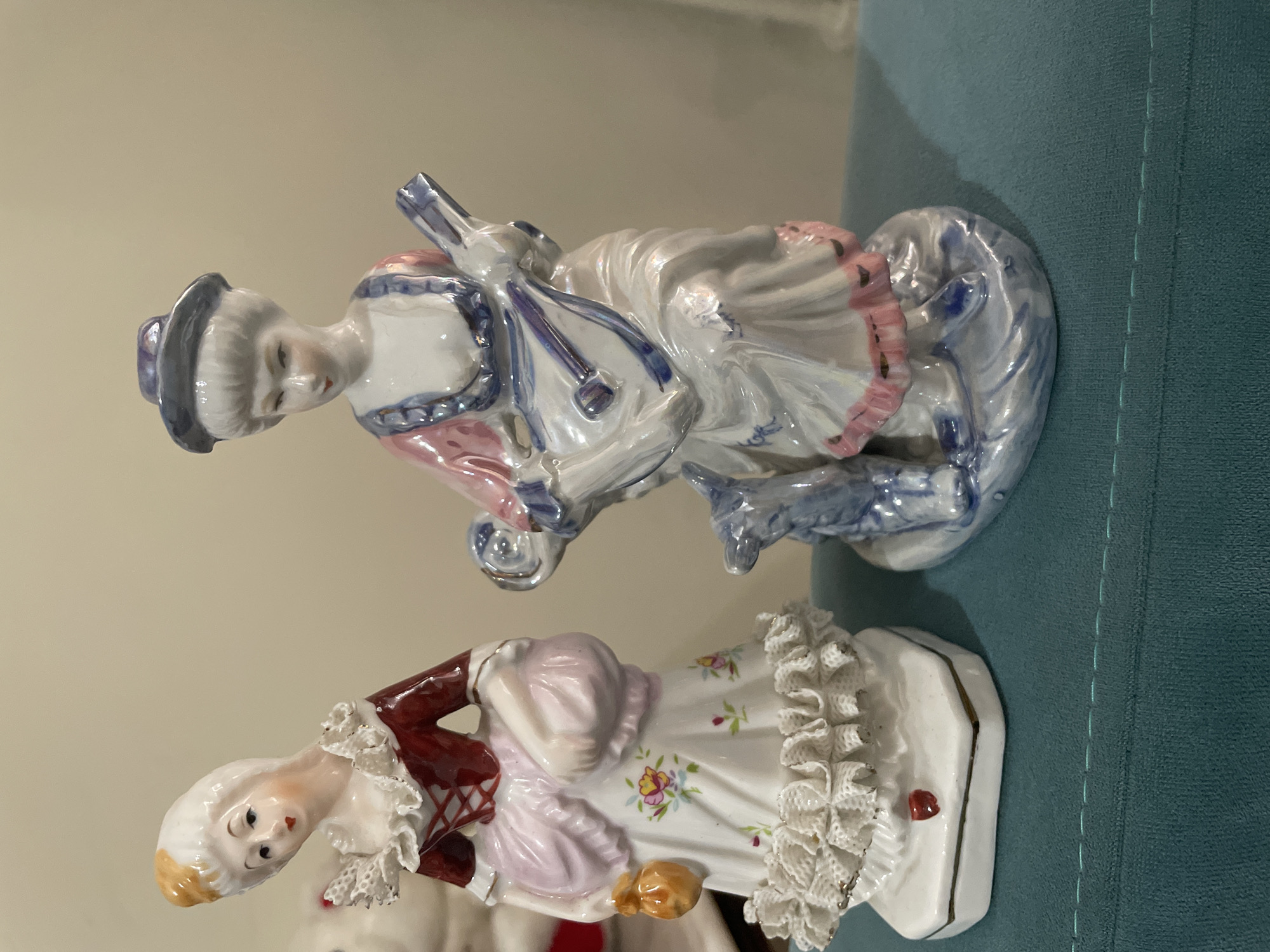 Two porcelain figurines of women in period clothing are displayed on a teal fabric surface. The figurine on the left is wearing a red bodice with white lace trim and a white skirt adorned with floral patterns and ruffles at the hem. She is holding a small orange pouch in her left hand and a light pink bag in her right. The figurine on the right is wearing a blue and pink dress and a blue hat. She is playing a stringed instrument, possibly a mandolin, and a small blue dog is at her feet. The background is a plain, light-colored wall. There is no indication of the location being in Kadıköy, Turkey.
